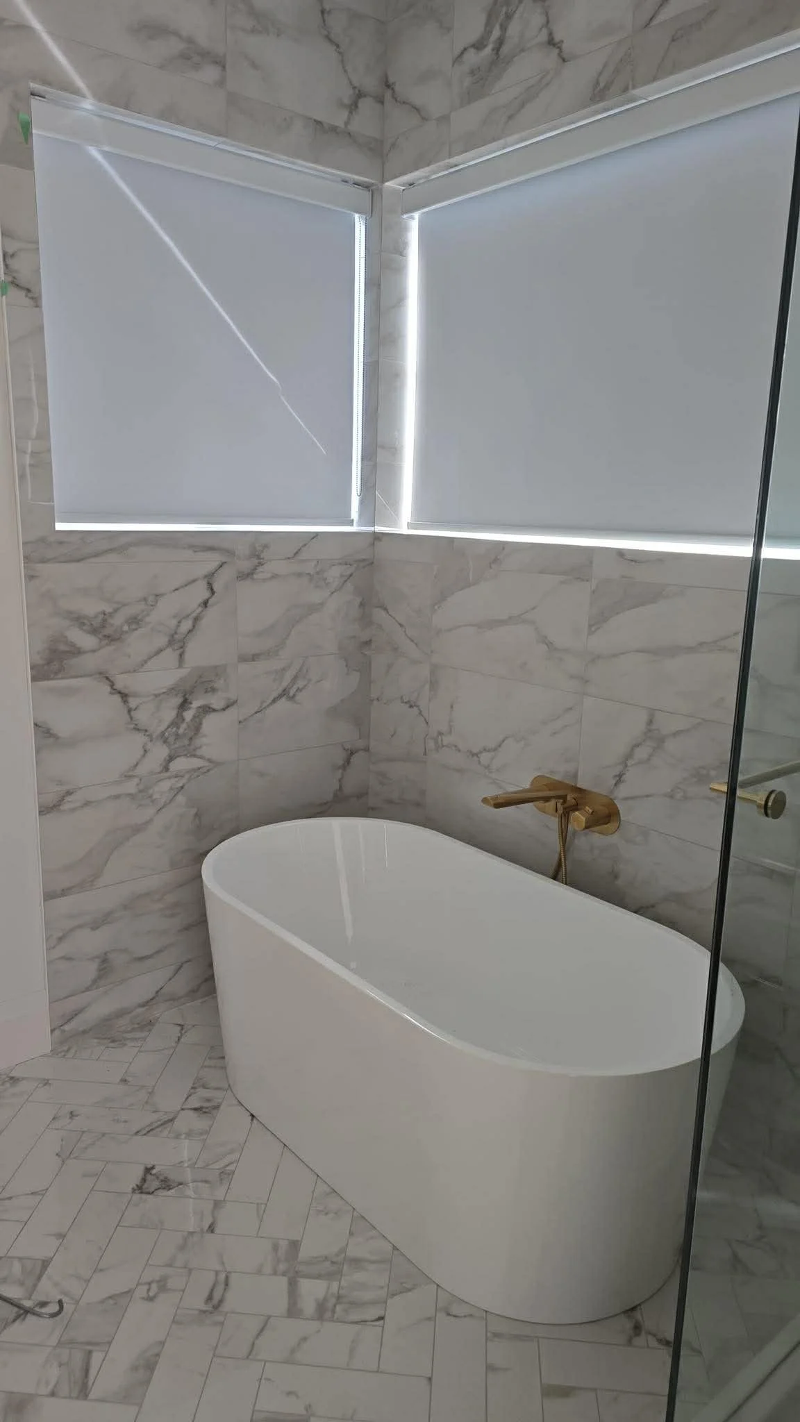 Modern bathroom with a freestanding white bathtub, marble tiled walls, and two frosted windows with sunlight streaming through.