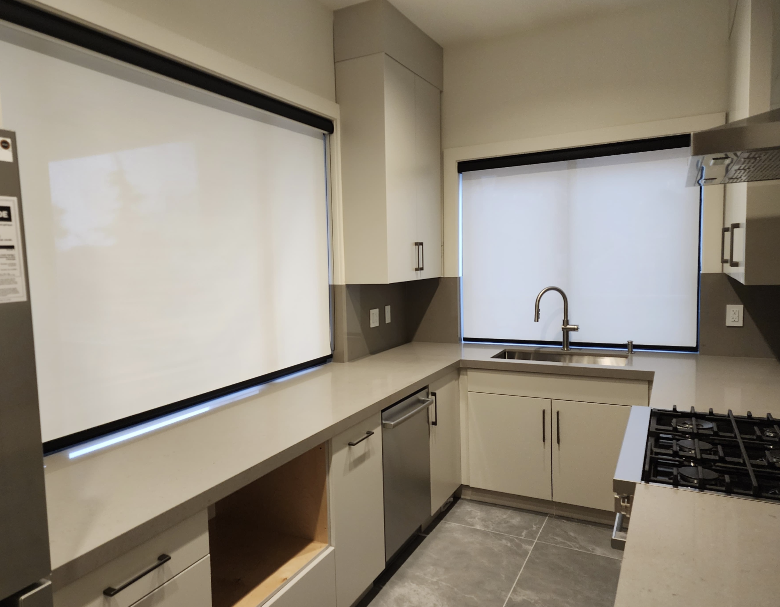 Modern kitchen with white cabinets, gray countertops, stainless steel appliances, and large windows with blinds.