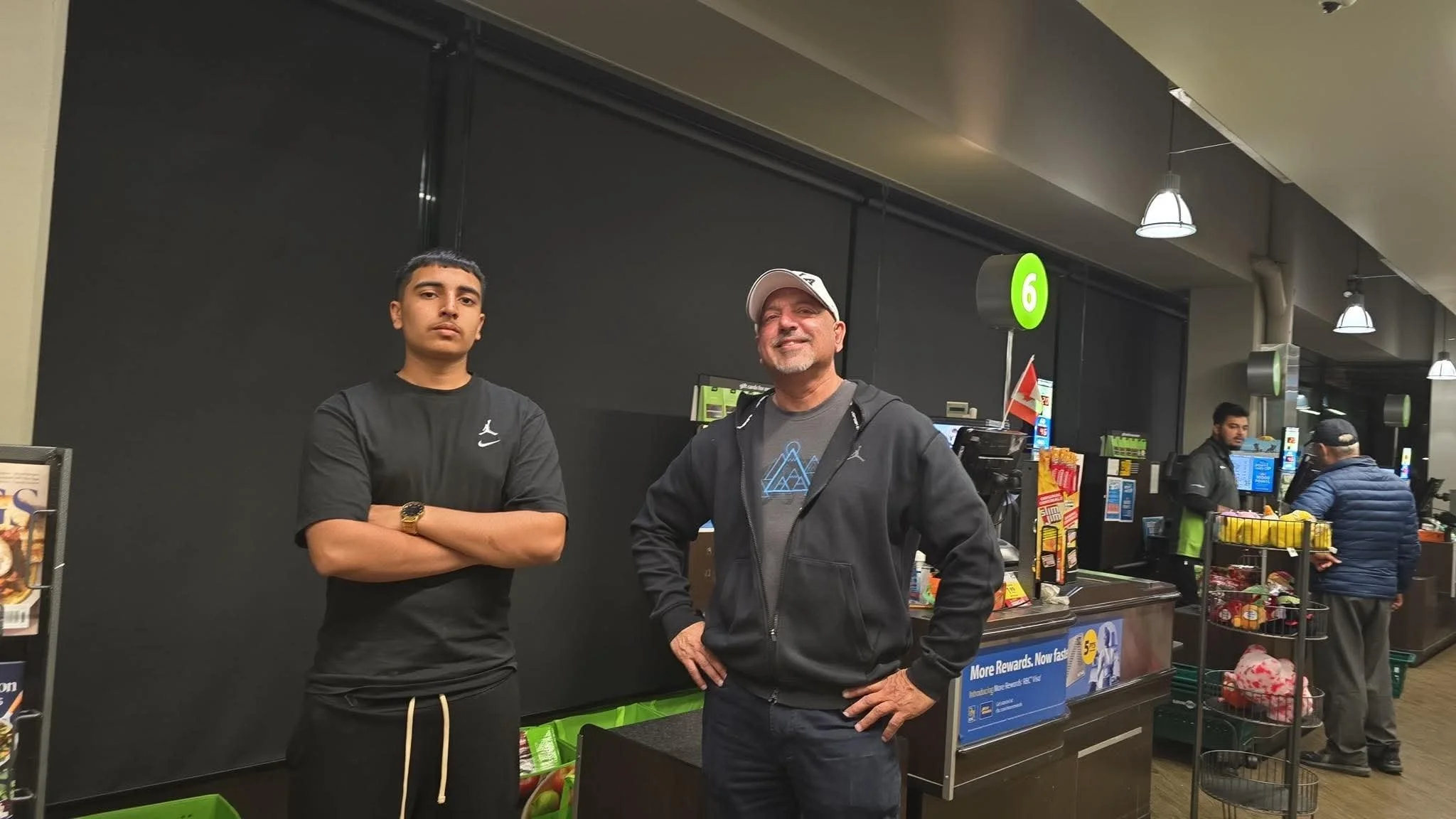 Two men standing inside a grocery store. The man on the left has short dark hair, crossed arms, and is wearing a black shirt and gold watch. The man on the right is smiling, has short hair, and is wearing a white cap, gray shirt, and black jacket. Store checkout counters and shoppers are visible in the background.