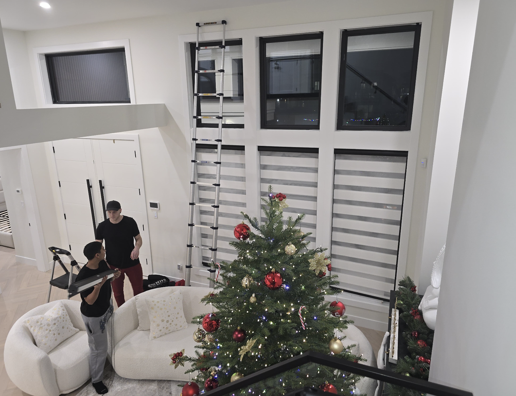 Living room decorated for Christmas with a decorated Christmas tree, a white curved sofa with gold pillows, a person holding a tray, another person standing nearby, a tall ladder against the wall, and large windows.