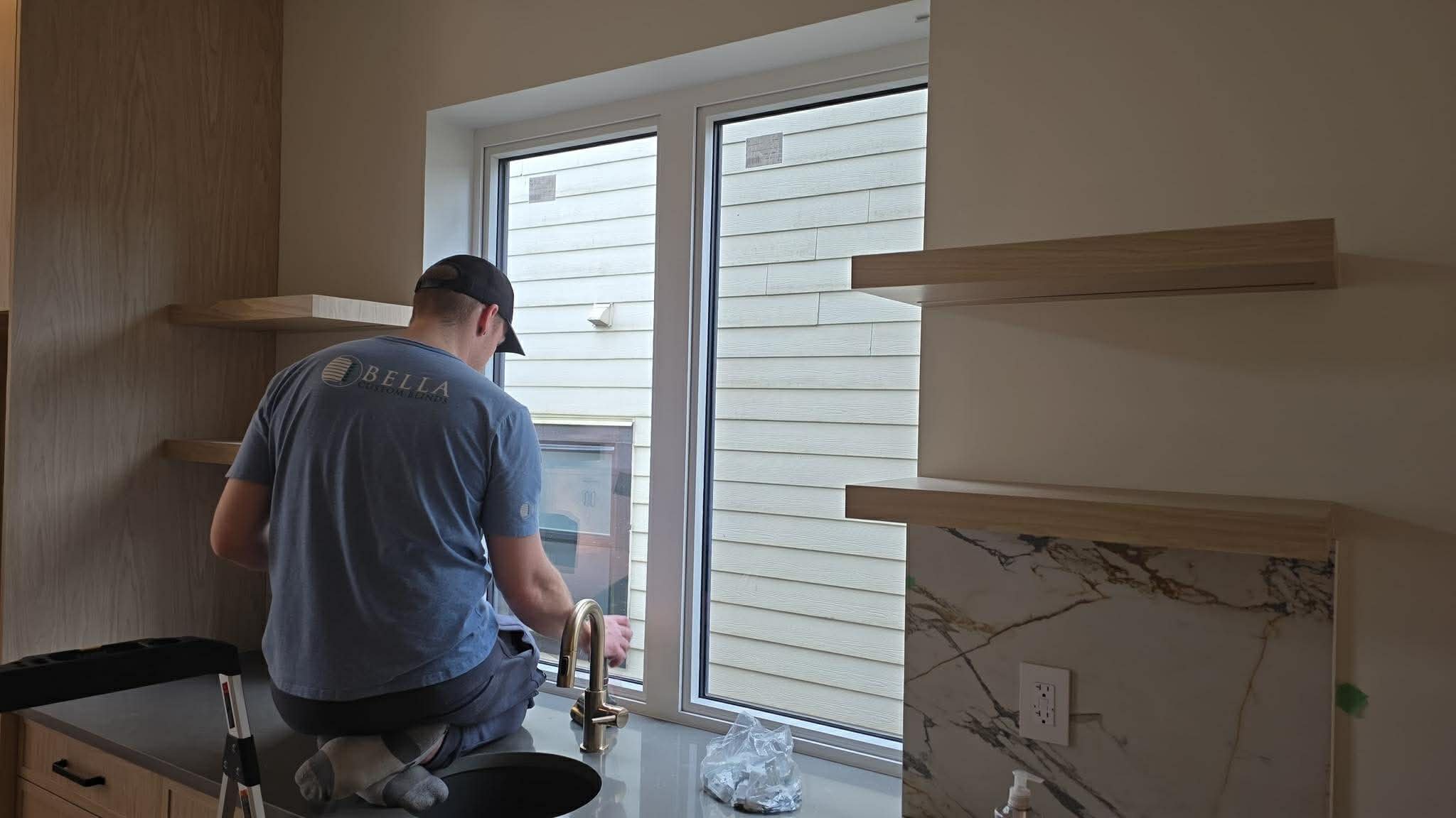 A person kneeling on a kitchen countertop, cleaning or examining a window, with wooden shelves and a marble backsplash visible in the kitchen.