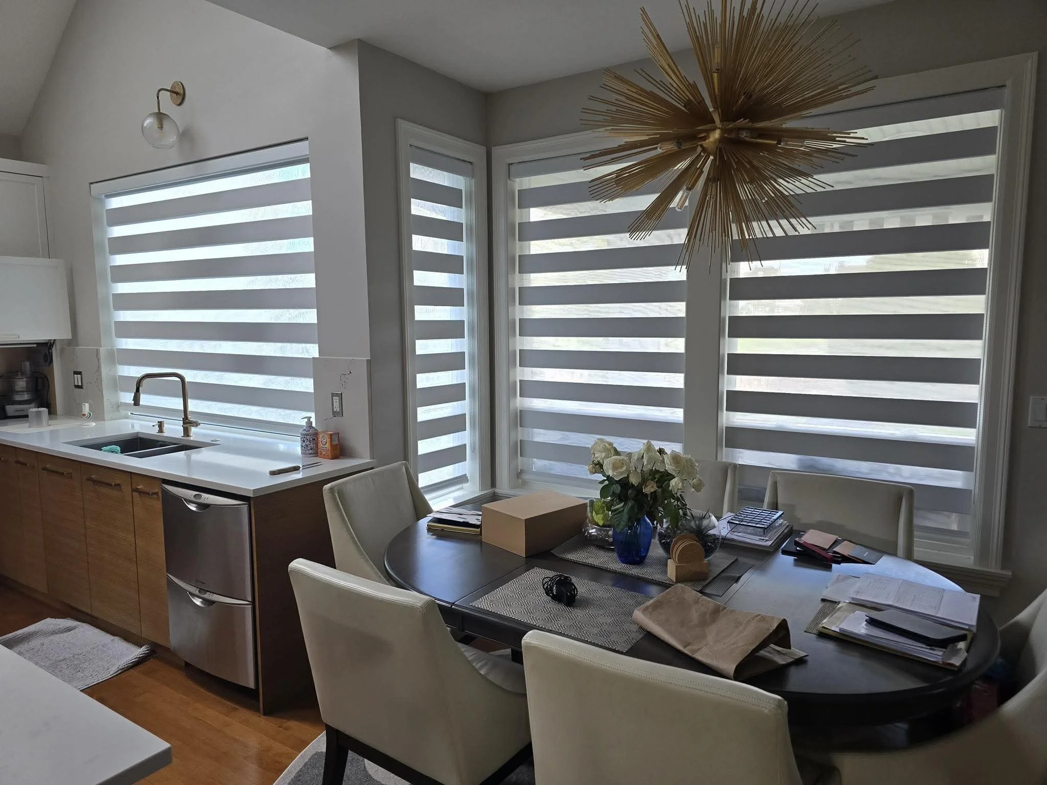 A dining area next to a kitchen with a window covering featuring horizontal blinds, a round table with white chairs, a vase of white roses, and various items on the table. There is a modern gold sunburst chandelier hanging above the table, and a window with similar horizontal blinds.