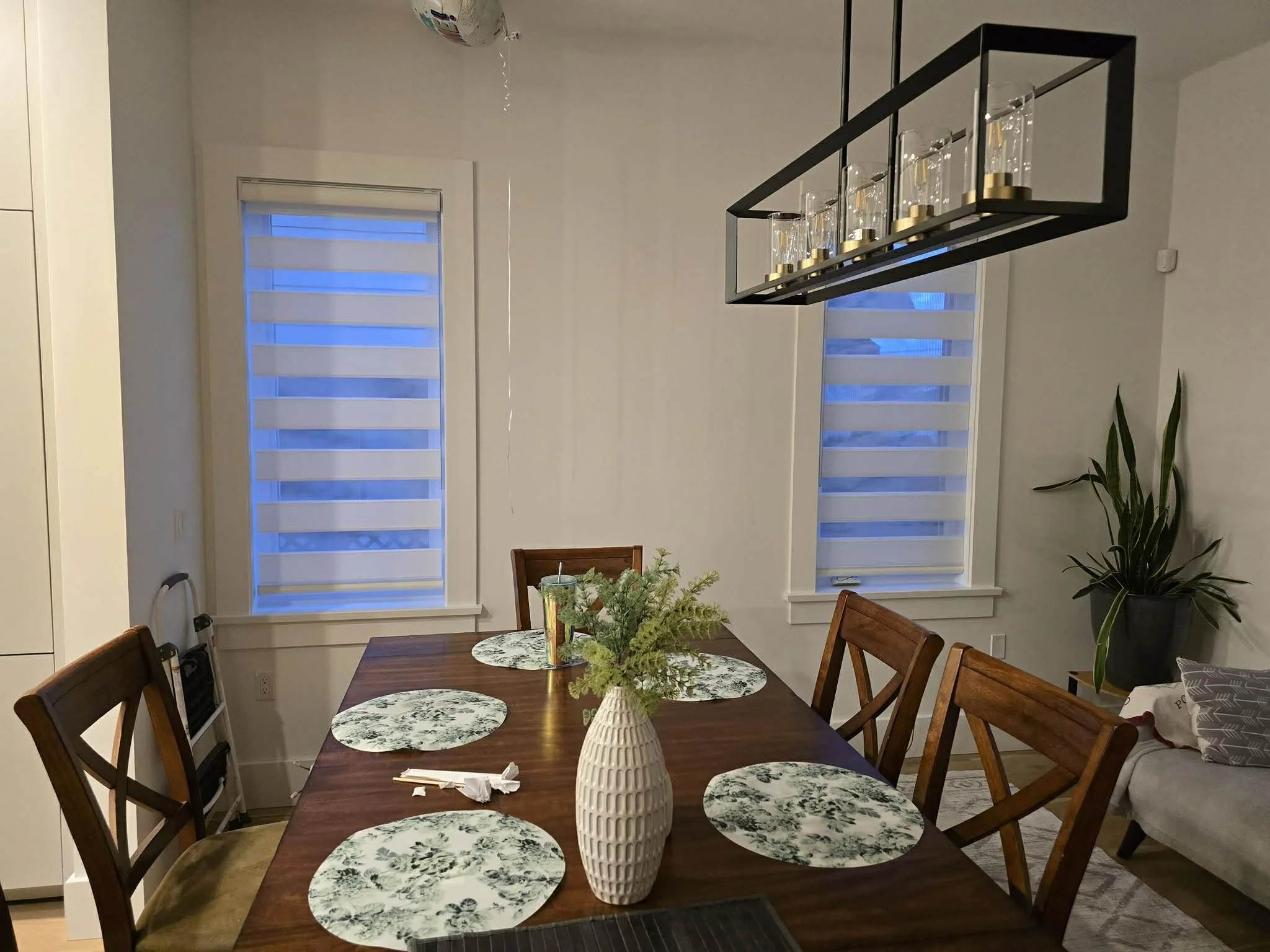 Dining room with a wooden table, six matching chairs, a white vase with greenery, four floral placemats, and windows with striped blinds