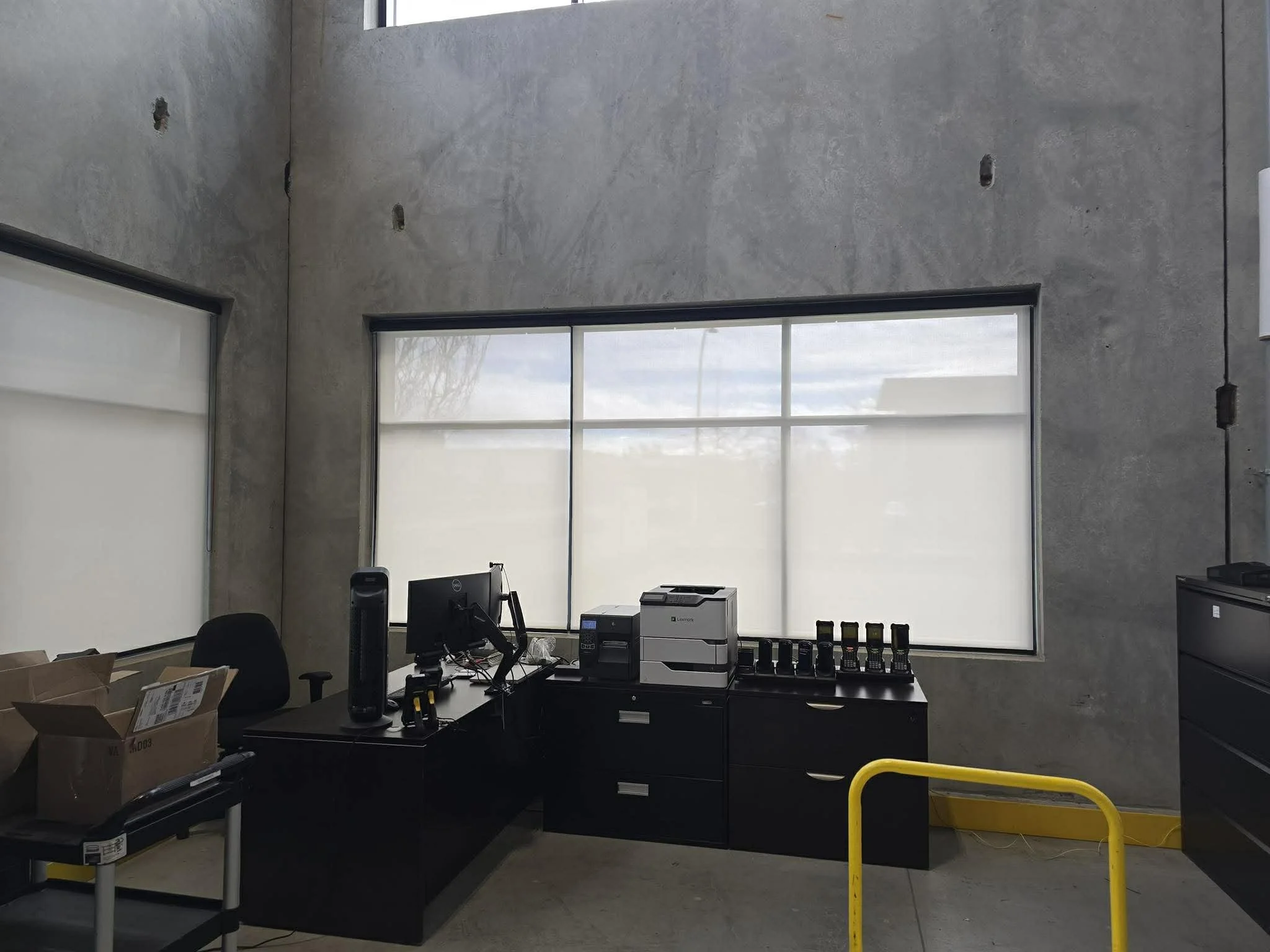 Empty office with a black desk, computer, monitor, and boxes, two large windows with shades, and unfinished concrete walls.
