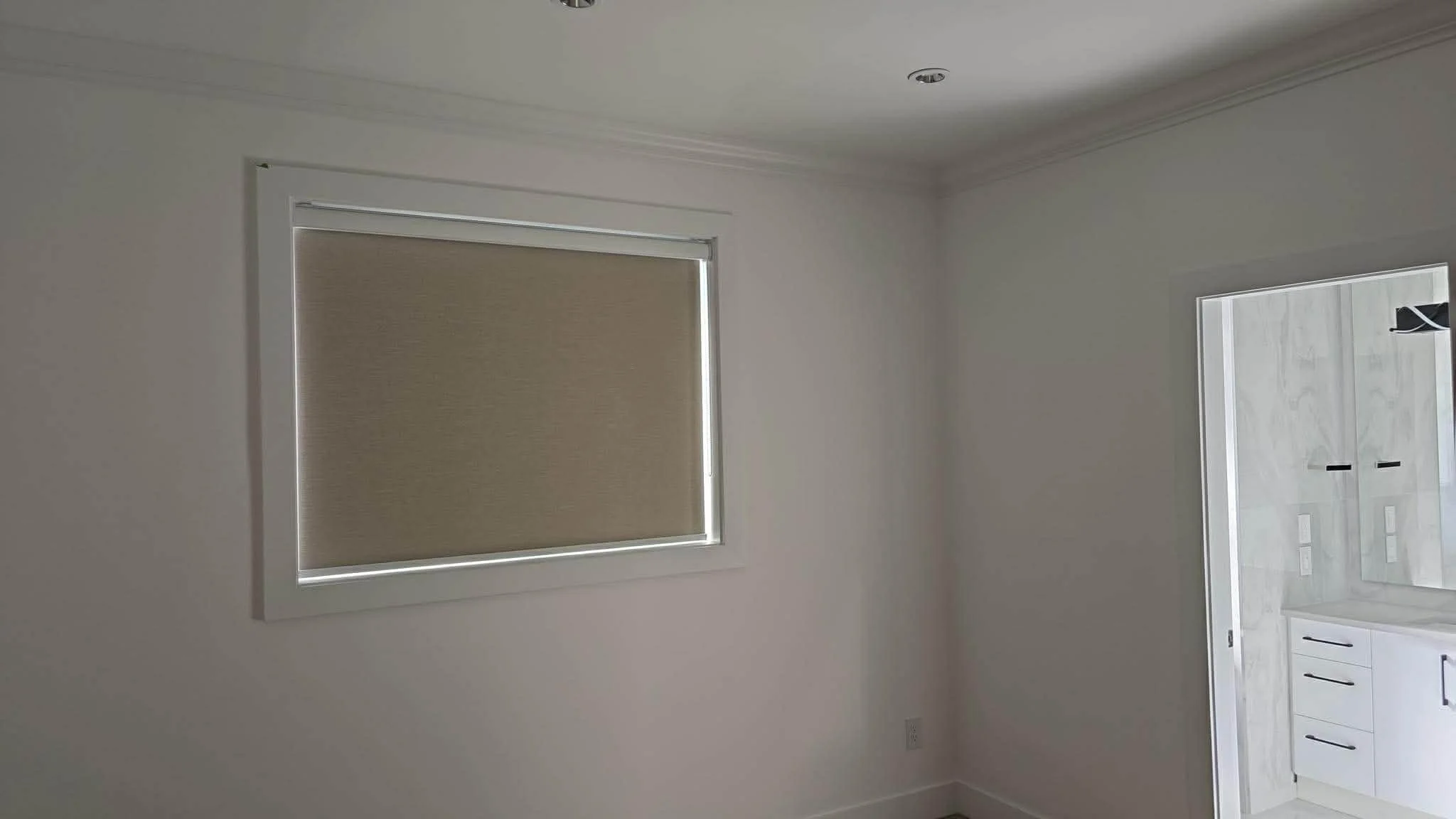 A room with a window covered by a beige roller blind, with a door opening into a bathroom with white cabinetry and marble walls.