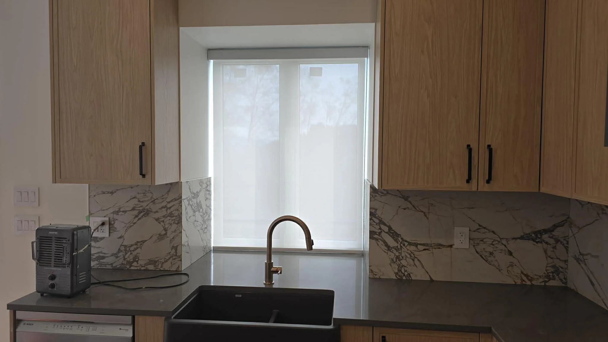 Modern kitchen space with wooden cabinets, a black countertop, a black sink, and a window with a white shade. There's a small black appliance plugged into an outlet on the left.