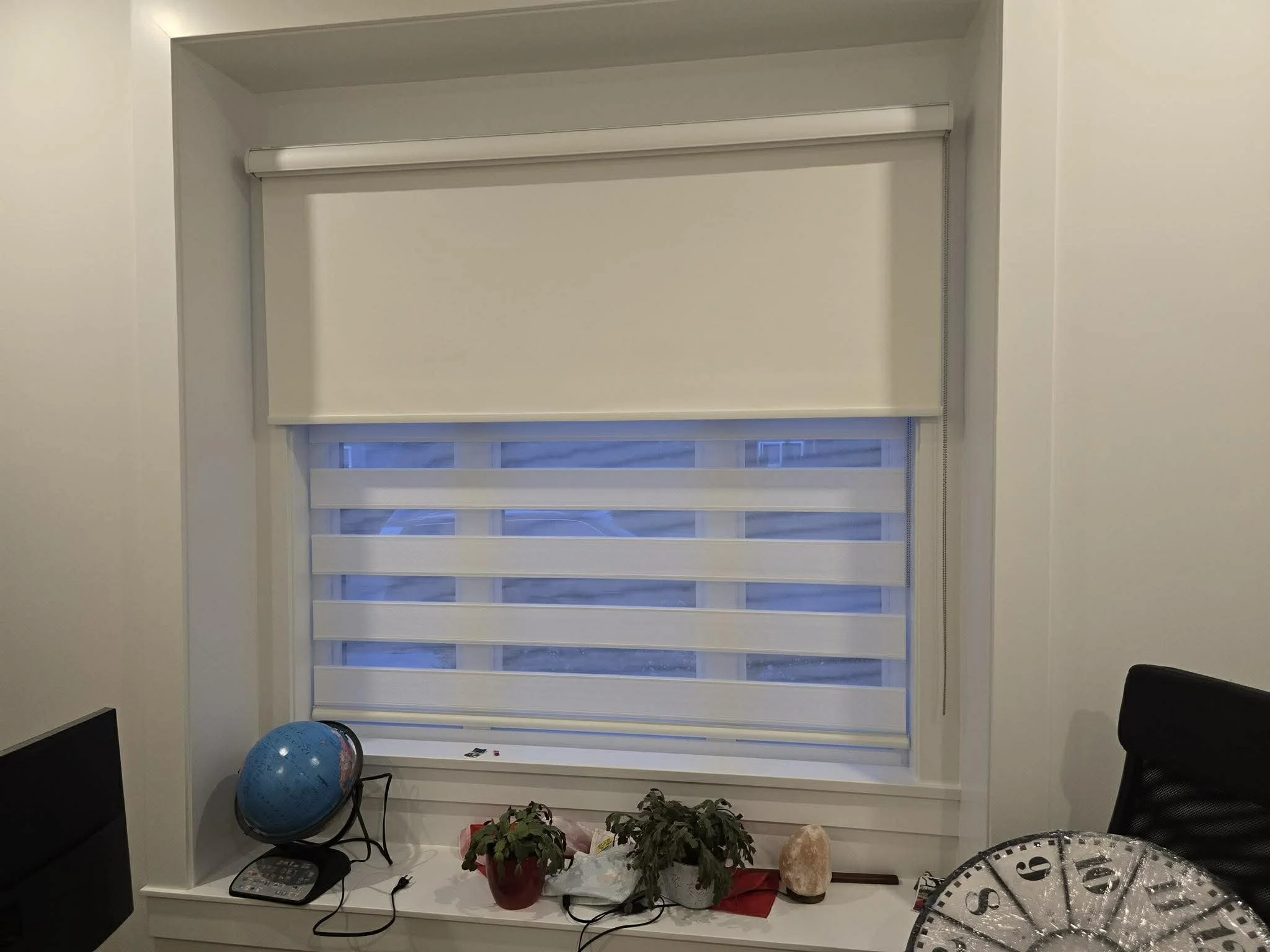 Office window with dual layer blinds, plants, a globe, and a salt lamp on the windowsill.