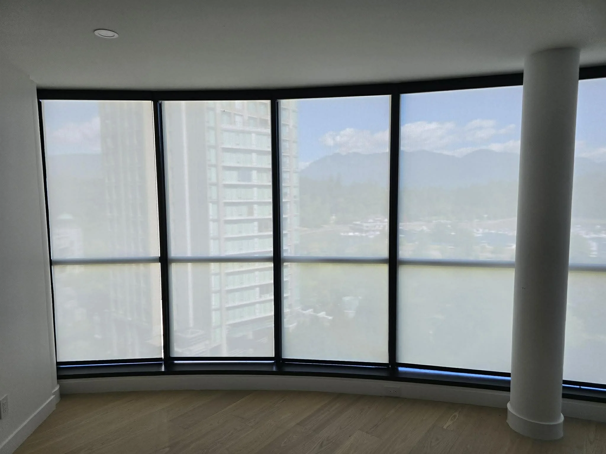 Empty room with large curved corner window overlooking city and mountains in distance.