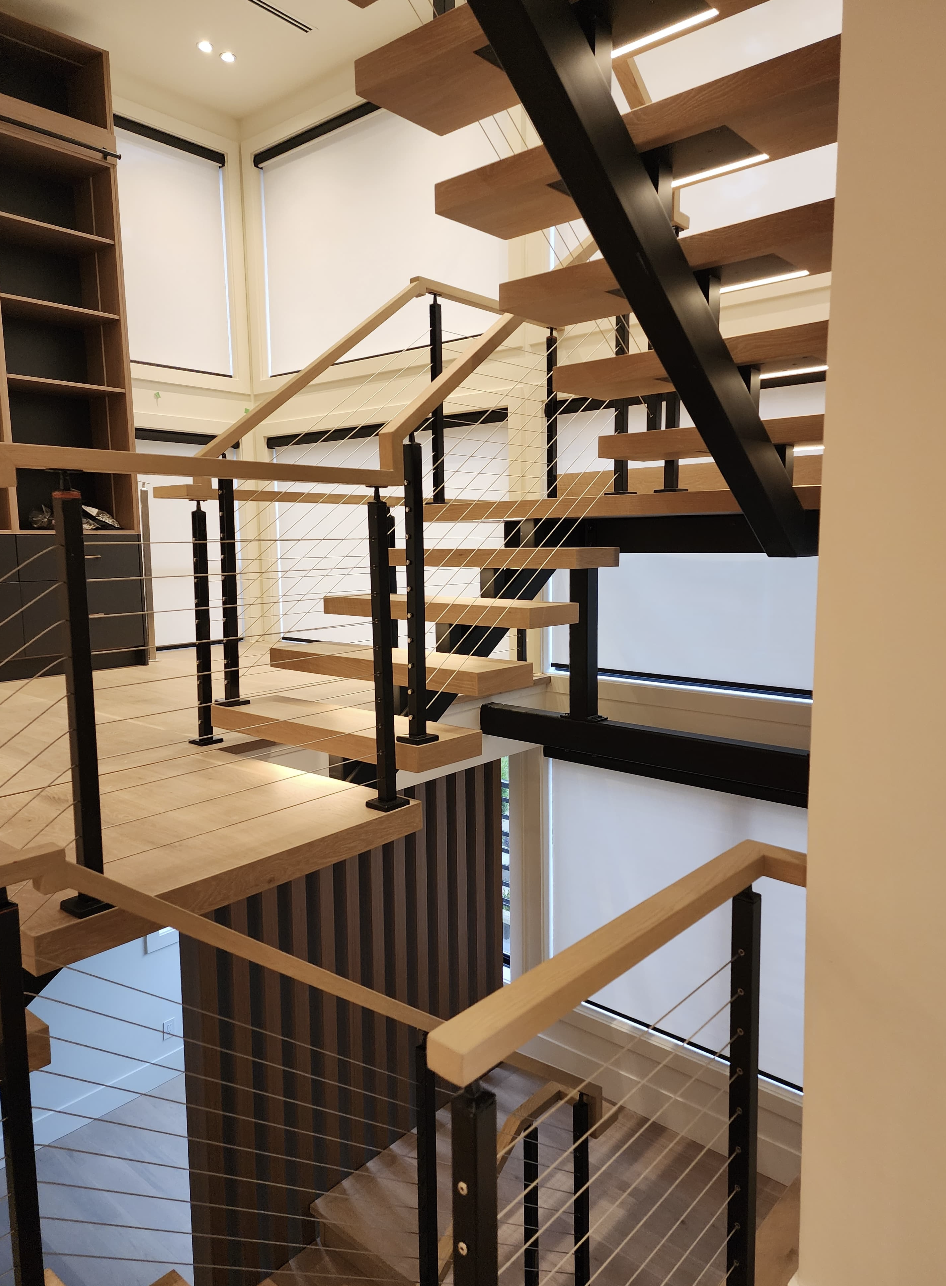 Modern indoor staircase with wooden steps, black metal supports, and thin cables serving as a railing, adjacent to windows with blinds.