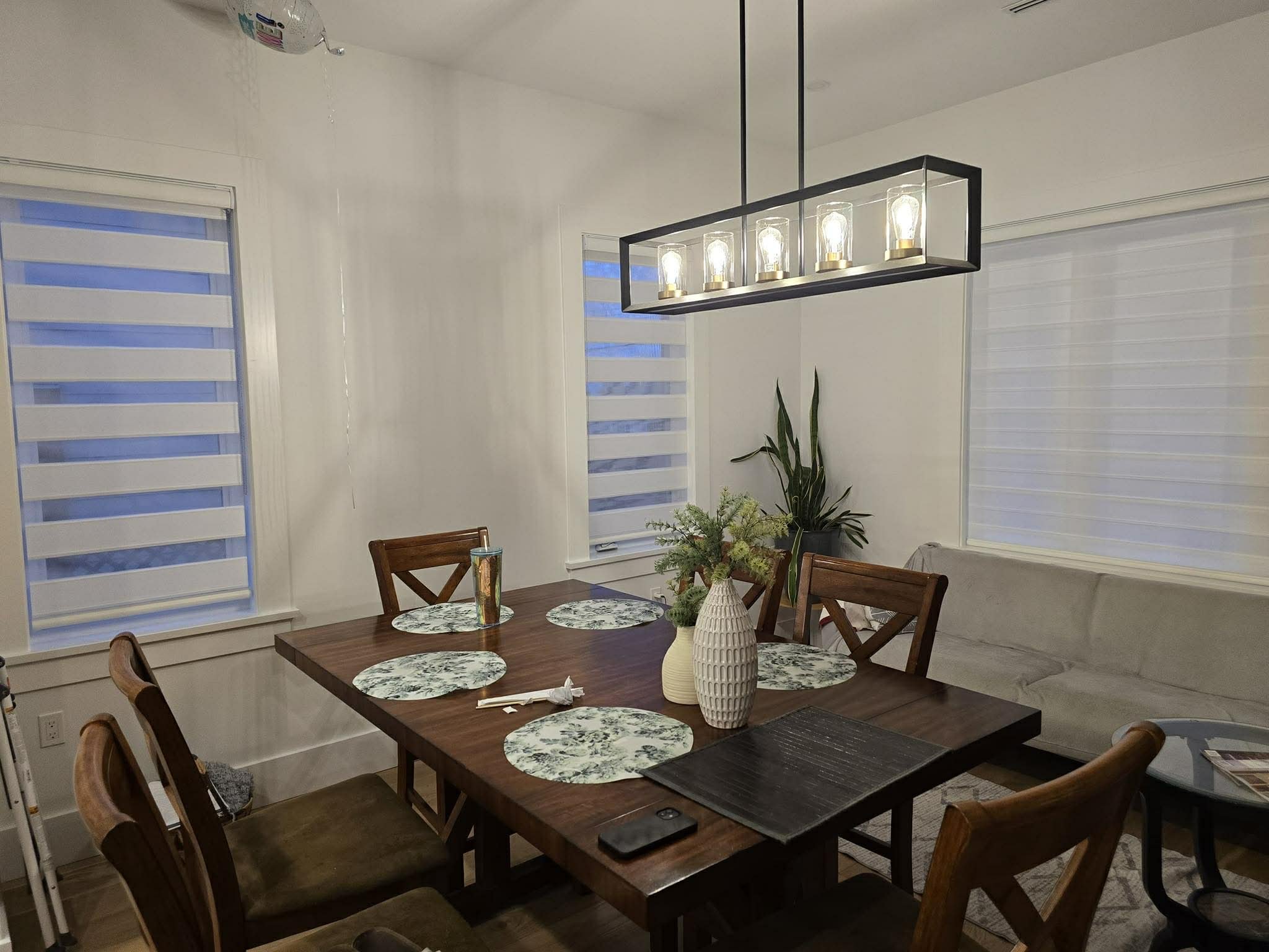 Dining room with wooden table, six chairs, white wall, windows with zebra blinds, hanging light fixture, sofa, potted plants, and various table settings and decor.