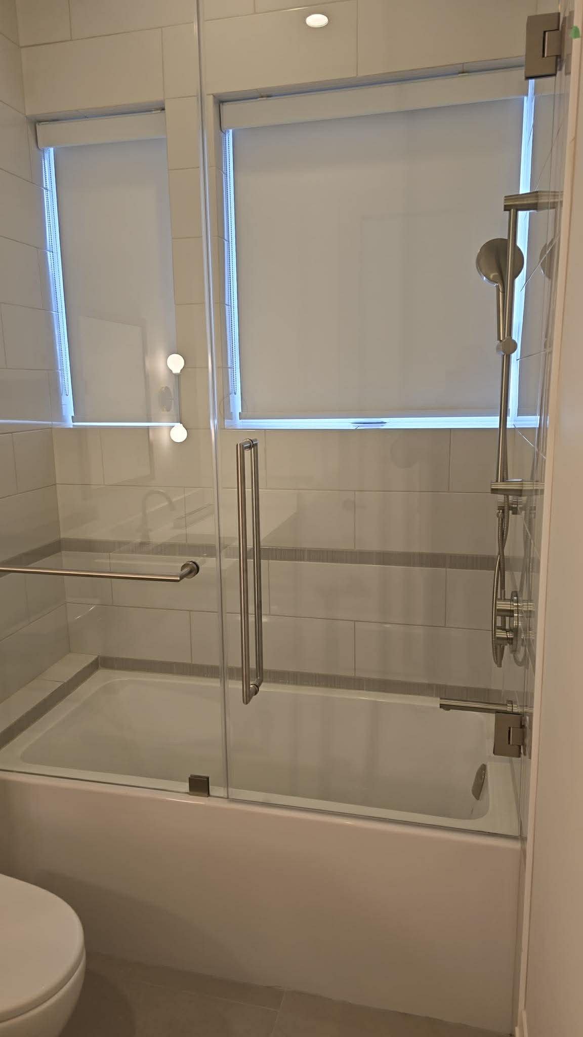 Bathroom with a glass shower enclosure, a window with blinds, and white tiled walls.