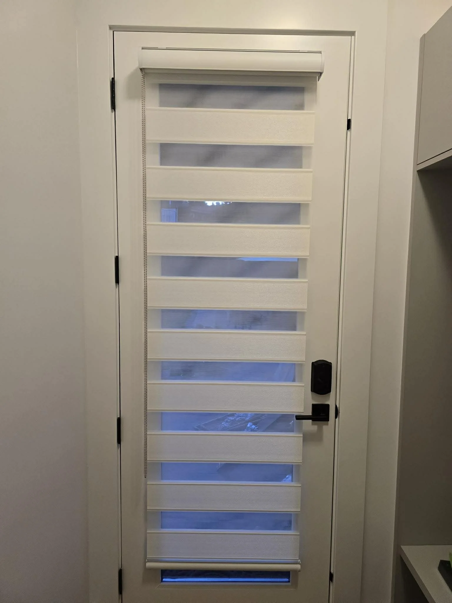 White exterior door with glass panels covered by a beige striped roller shade, black door handle, and smart lock.