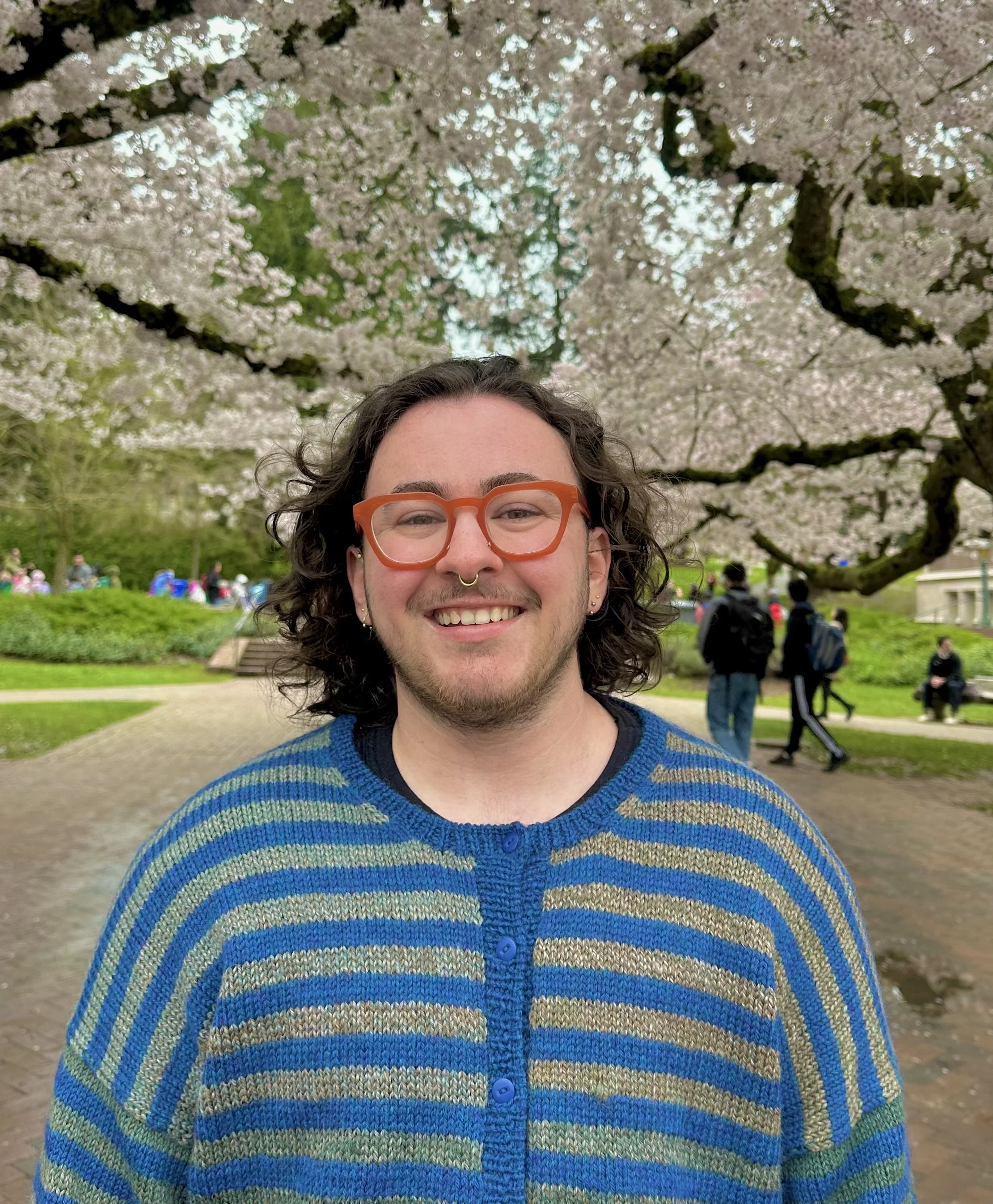 A smiling young man with curly hair, orange glasses, and a septum piercing stands outdoors in front of blooming cherry blossom trees during spring. People are walking and sitting in the background.