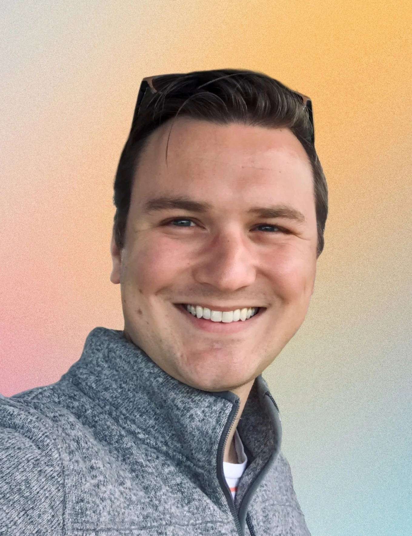 Close-up of a smiling person  with dark hair, wearing a gray zip-up jacket, standing in front of a gradient background with pastel colors.