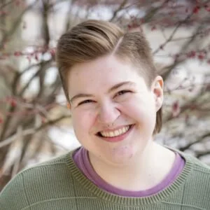 A smiling person with short light brown hair in a winter scene wearing a green sweater and purple t-shirt.