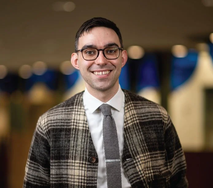 A smiling person with glasses, wearing a white collared shirt, gray tie, and plaid blazer, in a blurred indoor setting.