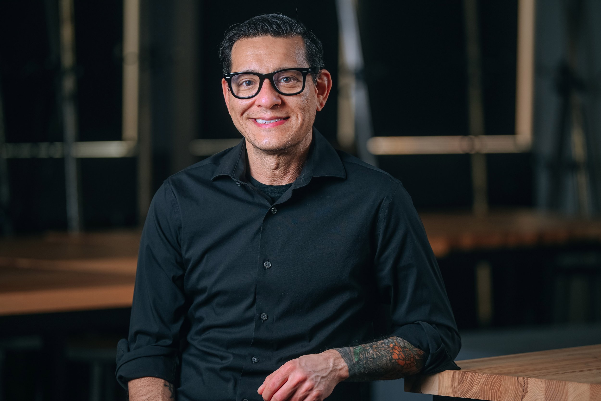 A smiling person with glasses wearing a black shirt, sitting at a wooden table in a studio setting.