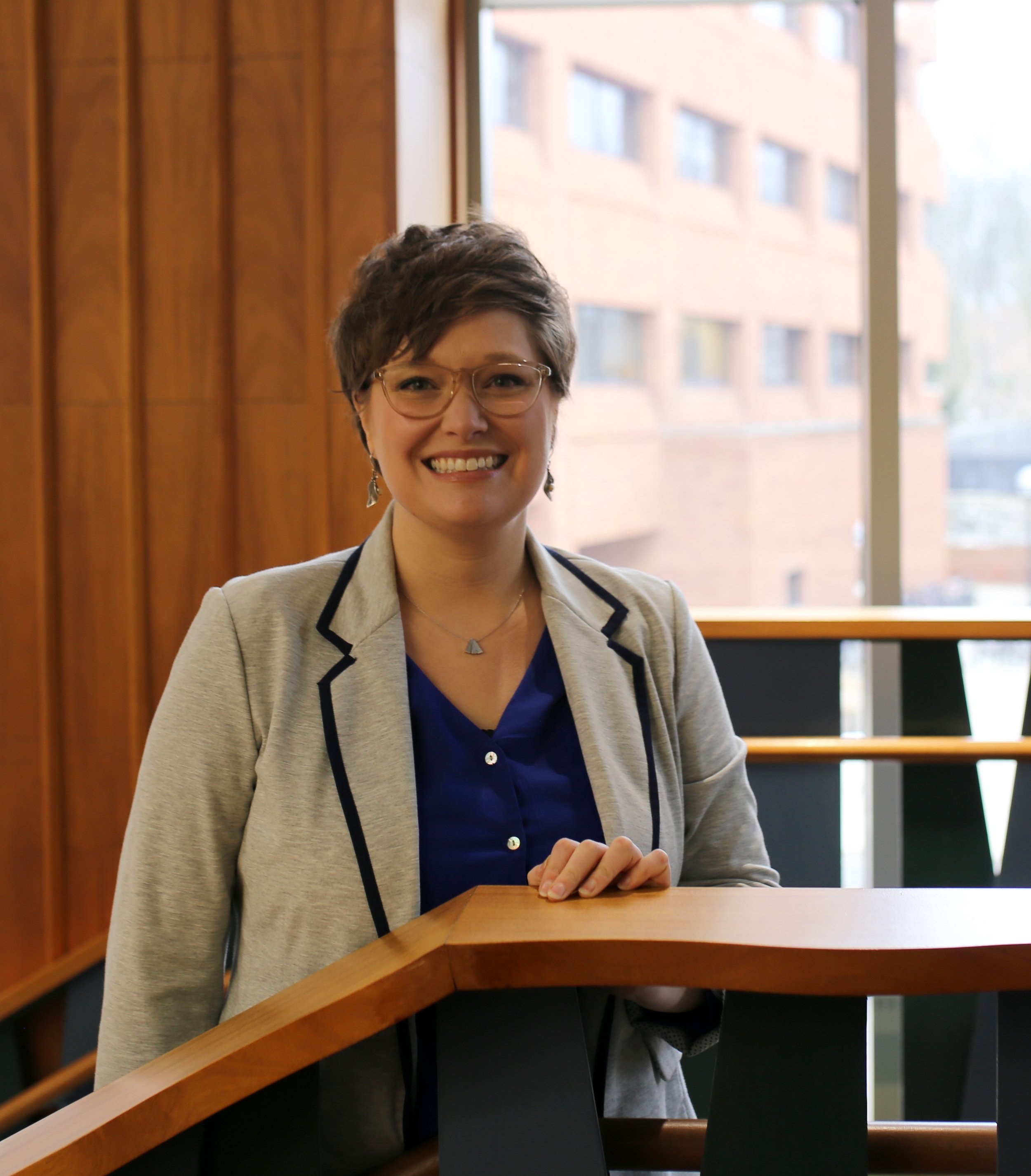 A smiling person with short hair and glasses, wearing a gray blazer and navy blouse, standing at a staircase near a wood-paneled window.