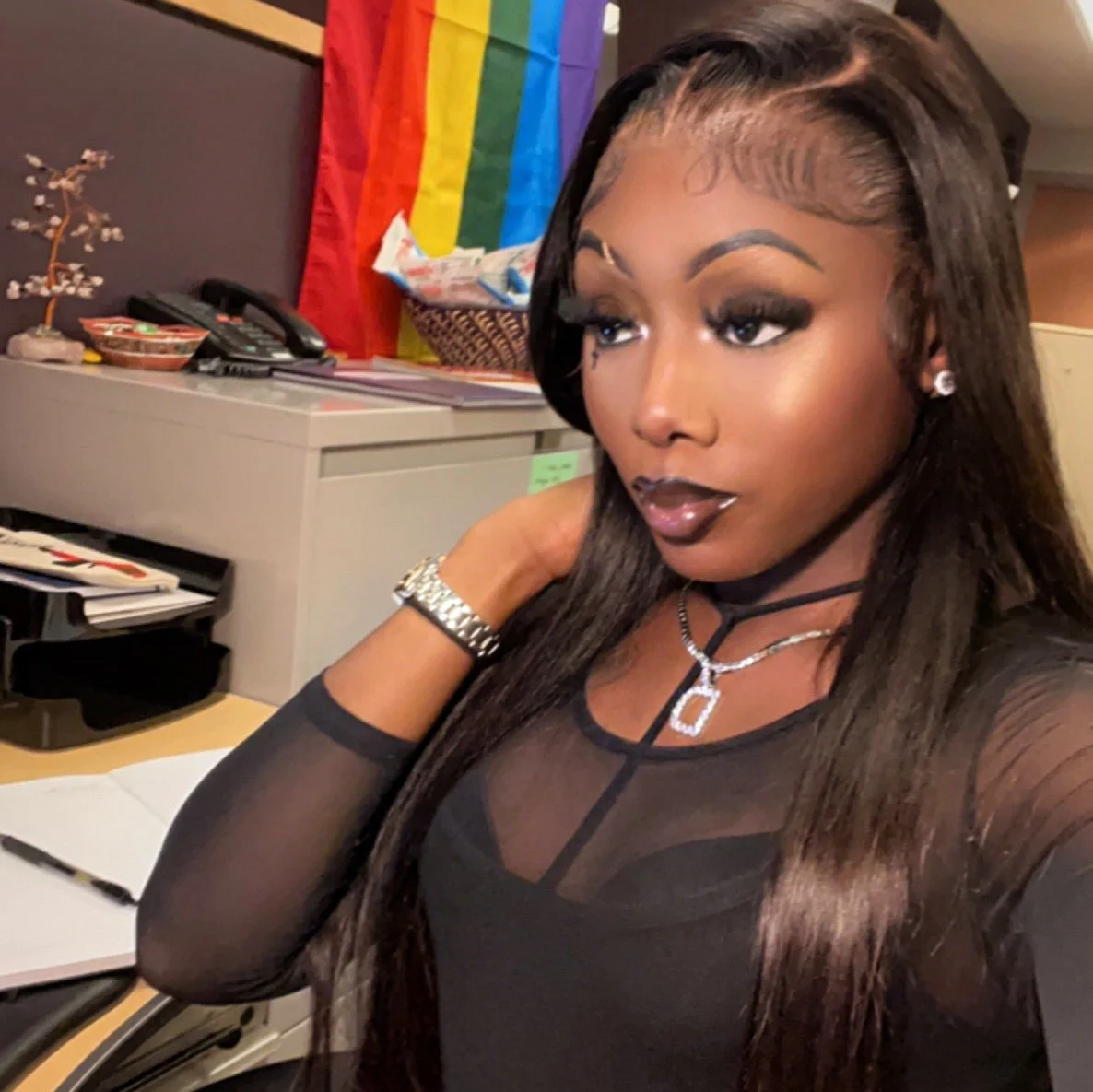 A smiling person with long with brown hair, wearing a black dress with silver jewelry sitting in an office..