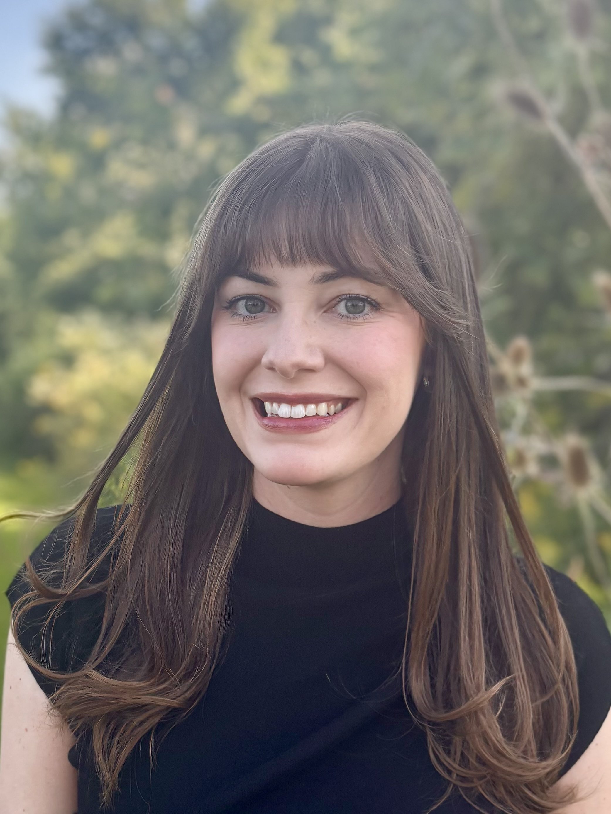 A smiling person with long brown hair and straight bangs, smiling outdoors with green trees in the background.