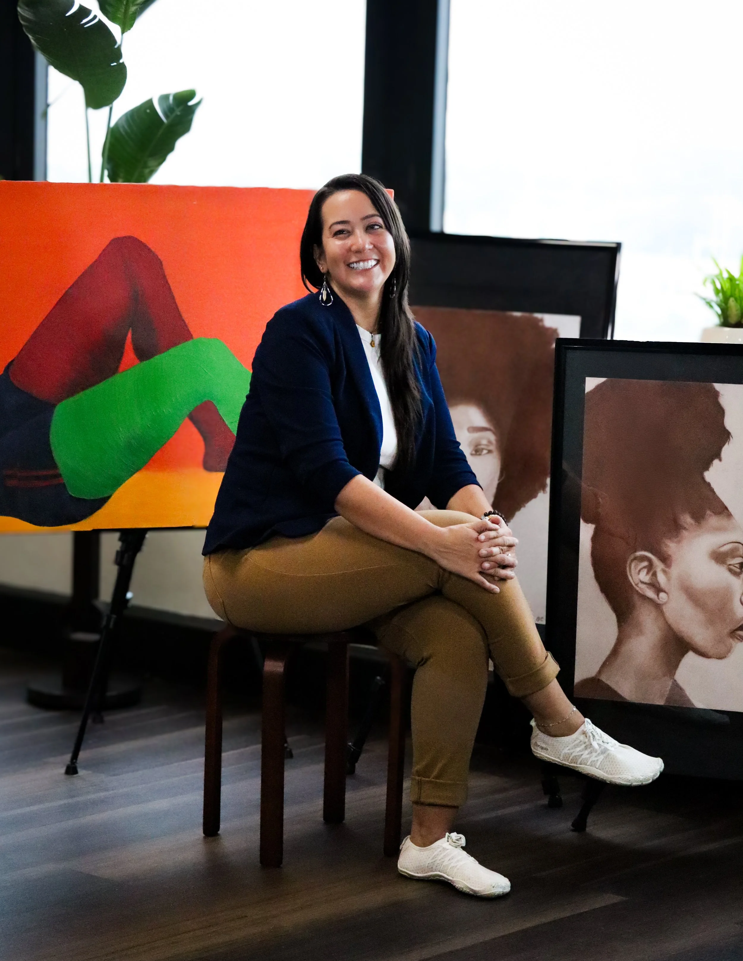 A woman with long dark hair sits on a wooden stool, smiling at the camera, in an art gallery with paintings of African American women and a red couch with green and black artwork behind her.