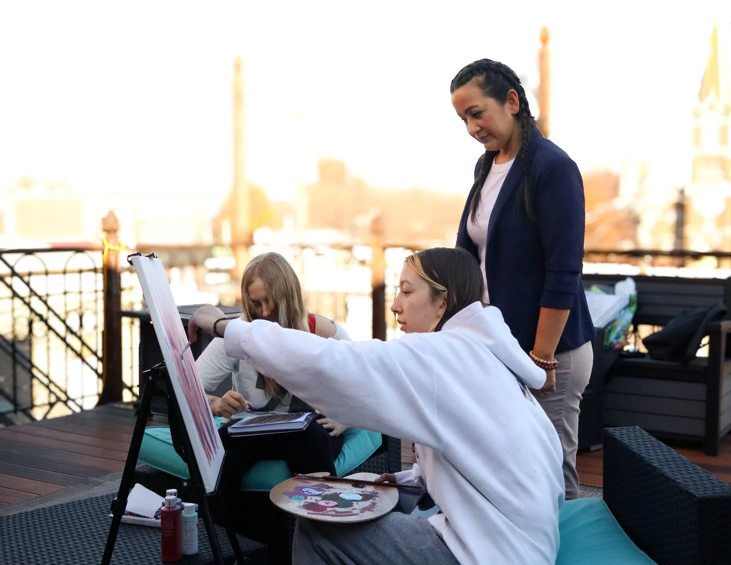 Three women painting outdoors on a deck, with one standing and two seated, using an easel and art supplies, with a city skyline in the background.