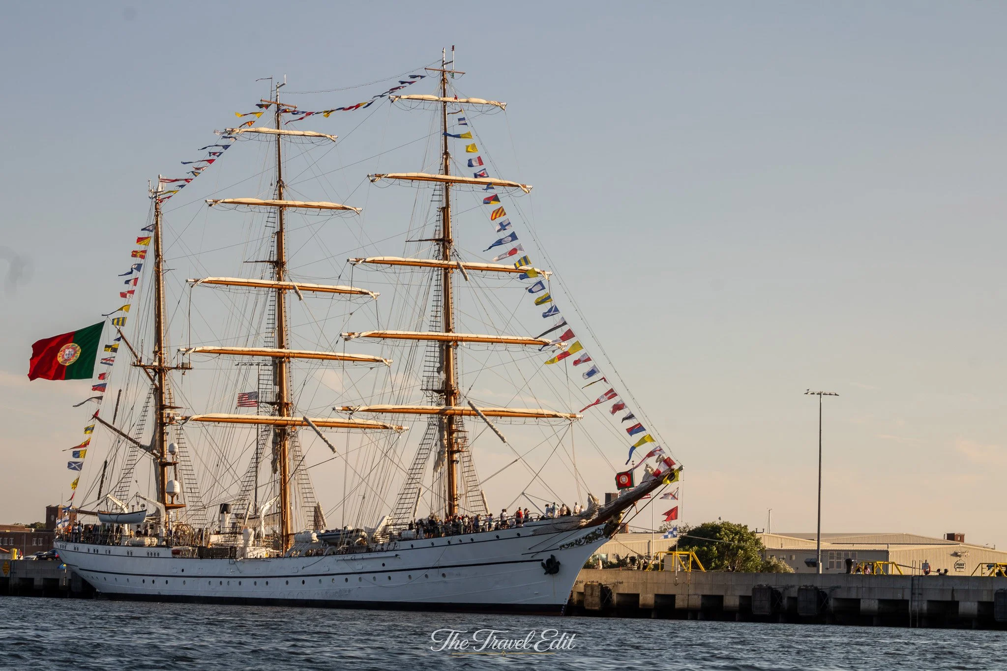 A large sailing ship with multiple masts is docked at a harbor. The ship is decorated with numerous colorful signal flags, and the American and Portuguese flags are visible. People are seen on the deck and the dock in the background, with a clear sky overhead.