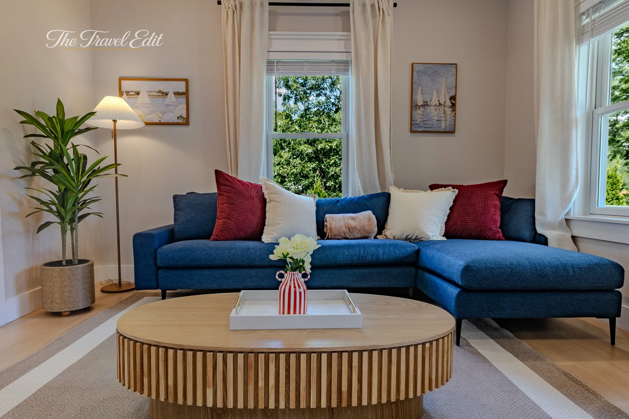 Living room with a blue sofa, red and white pillows, a wooden coffee table with a striped vase of white flowers, a potted plant, a tall floor lamp, and framed artwork of sailboats on the walls; large windows with cream curtains showing greenery outsi