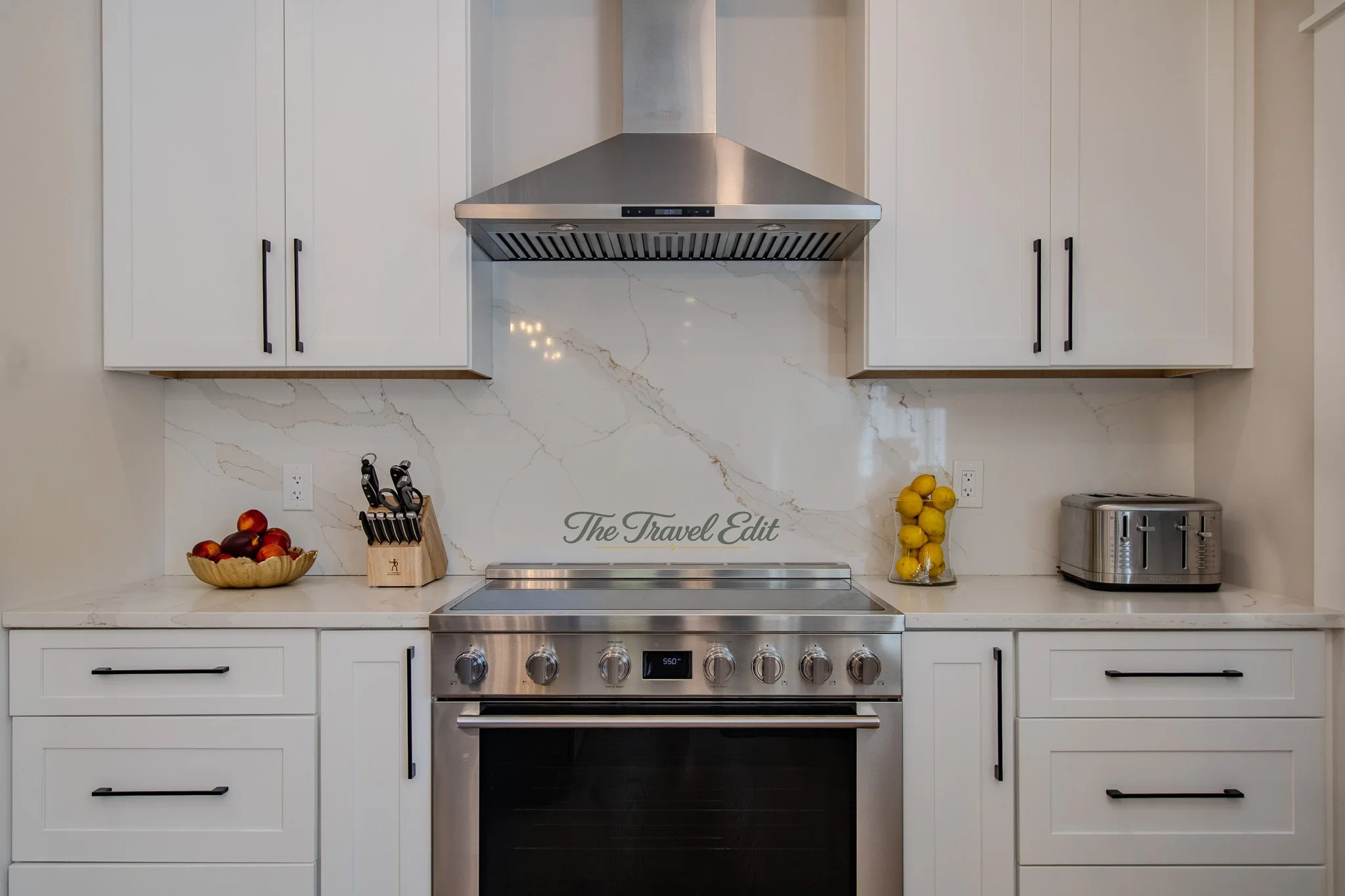 Kitchen with white cabinets, marble backsplash, stainless steel stove, and kitchen appliances including a toaster. Decorative items include apples in a bowl and lemons in a glass jar.