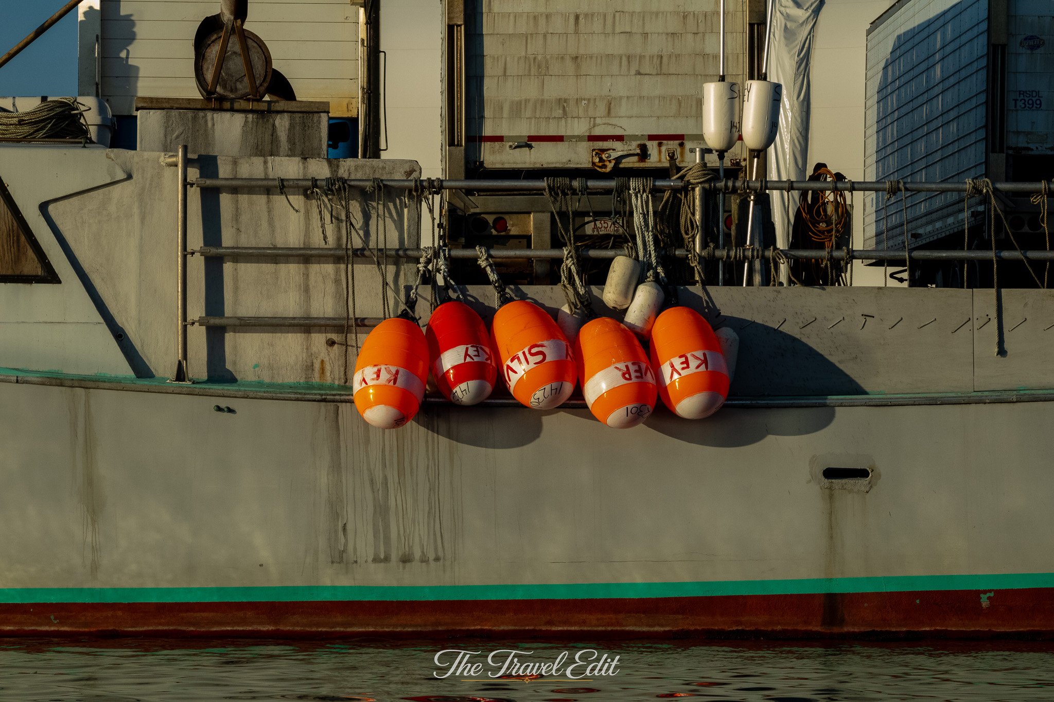 Close-up of orange and white buoy floats with black markings, tied to a metal railing on dockside vessel, with various equipment and storage containers in the background.