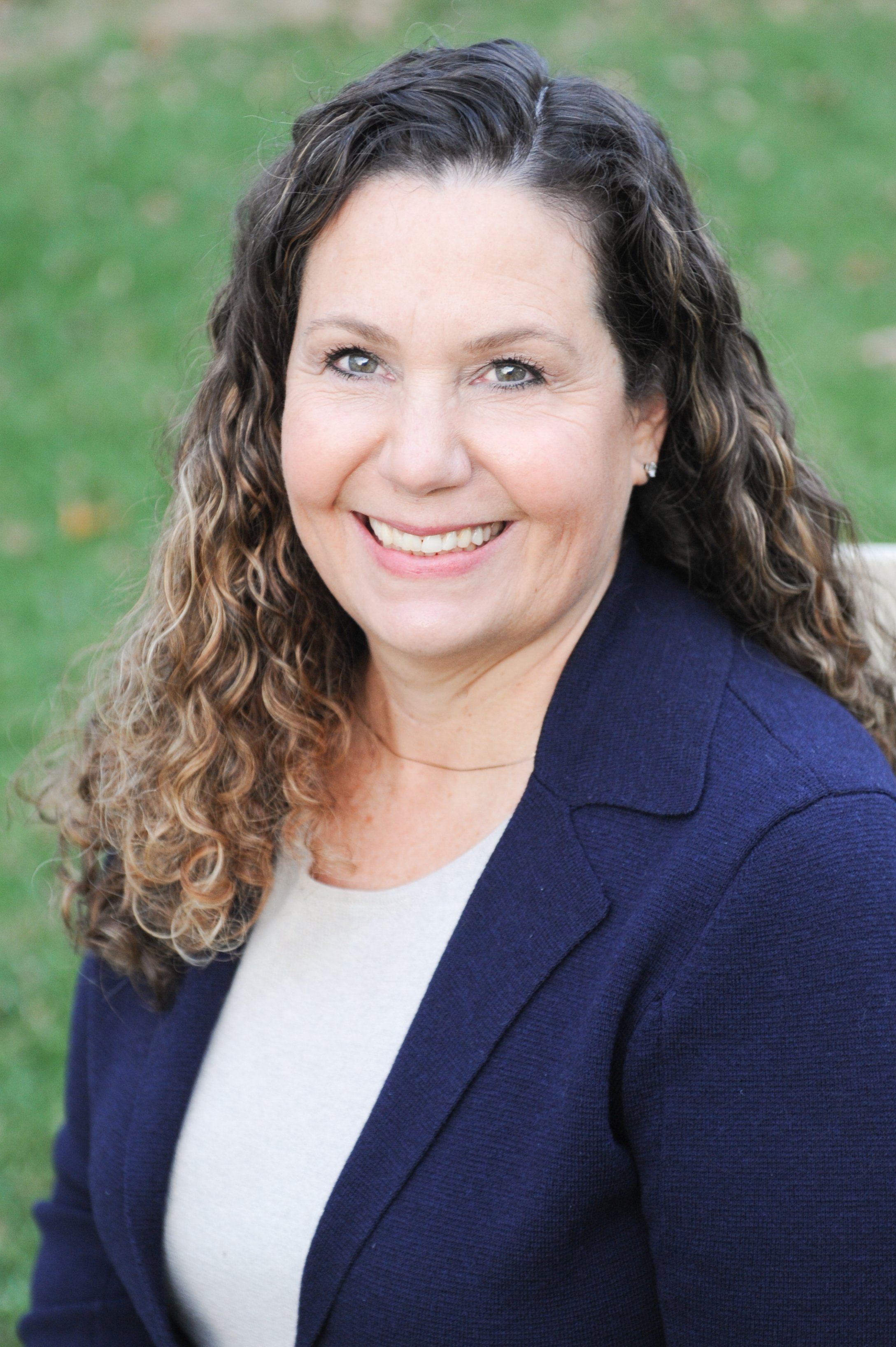 A smiling woman with curly brown hair, wearing a navy blazer and light top, outside on green grass.