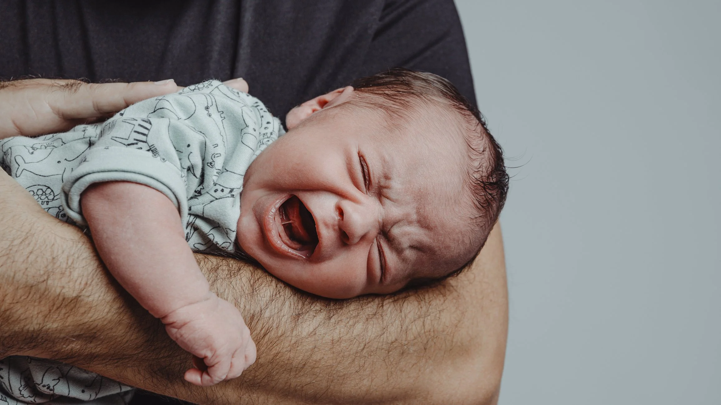Close-up of a yawning newborn baby wrapped in a white blanket with a striped blue, pink, and white background.