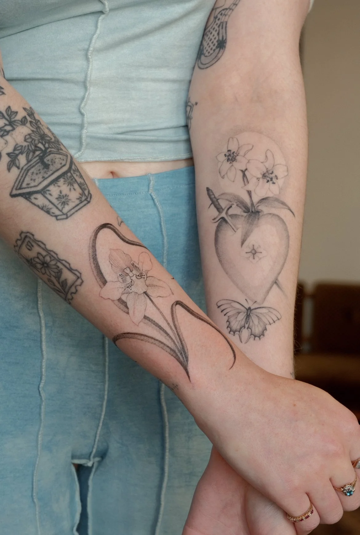 Close-up of person's arms with various black and gray tattoos, including a potted plant, flowers, and a heart with a butterfly below, against a background of light clothing. One tattoo is a sacred heart and the other one a fineline orchid.