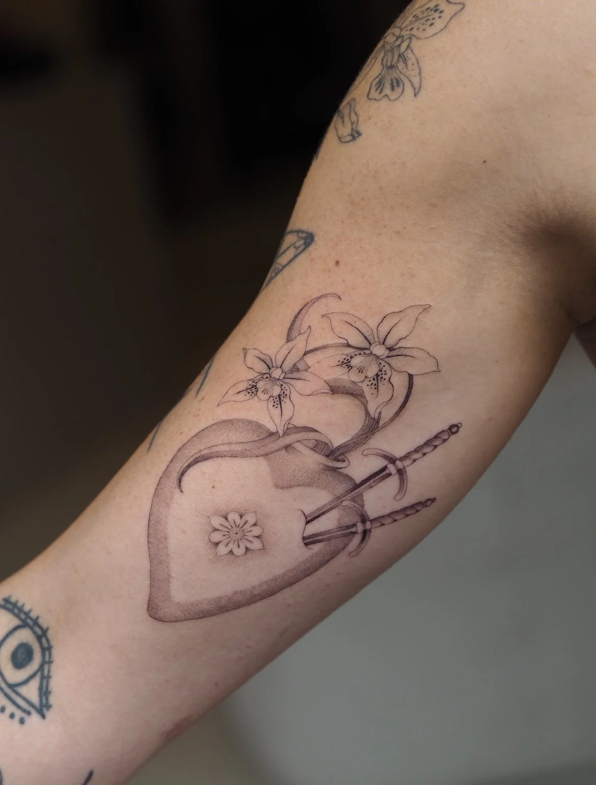 Tattoo of a sacred heart with orchids and a sword on a person's arm.