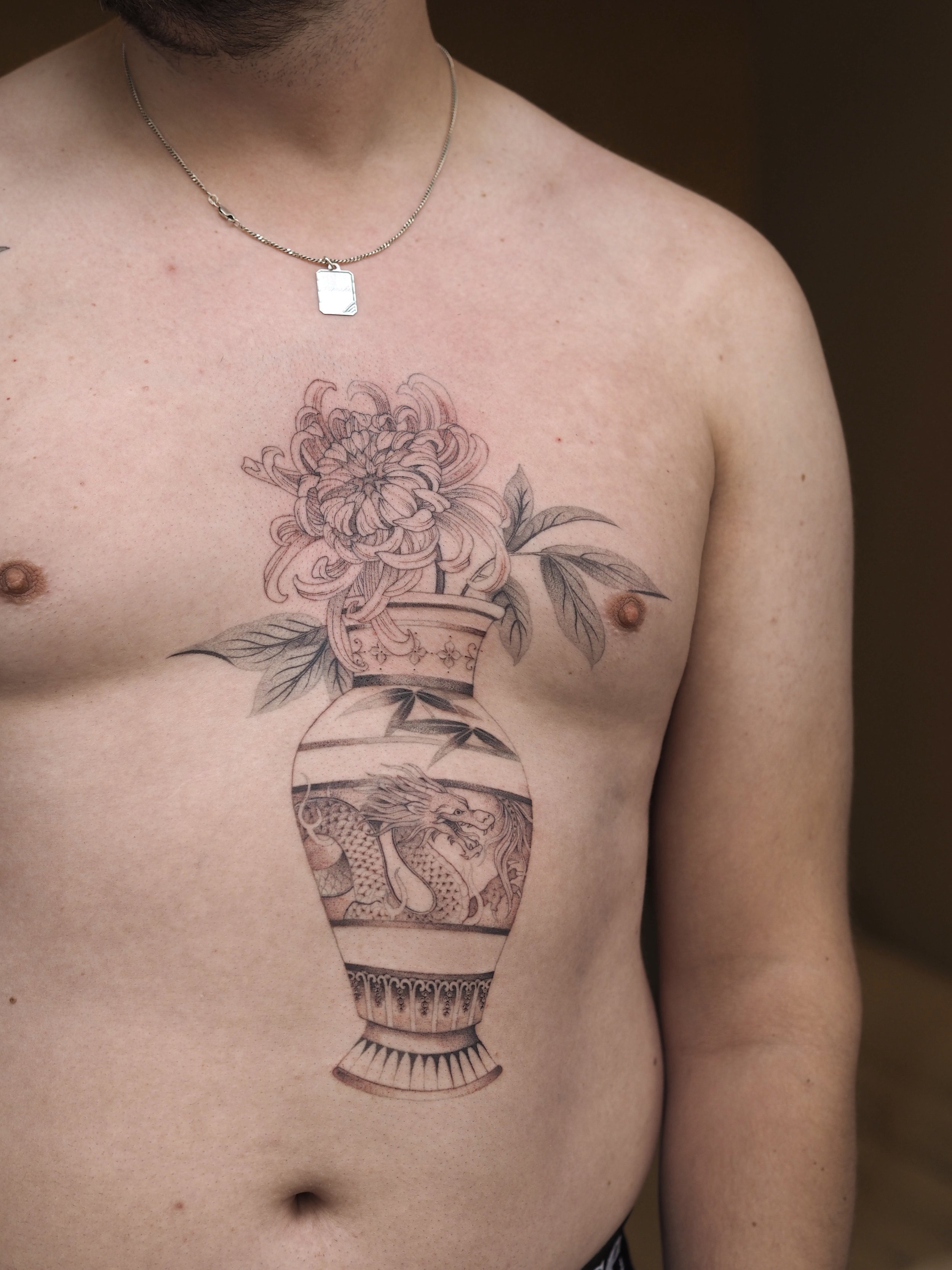 Tattoo of a vase with a dragon and chrysantemum flowers on man's chest, wearing a silver chain necklace.