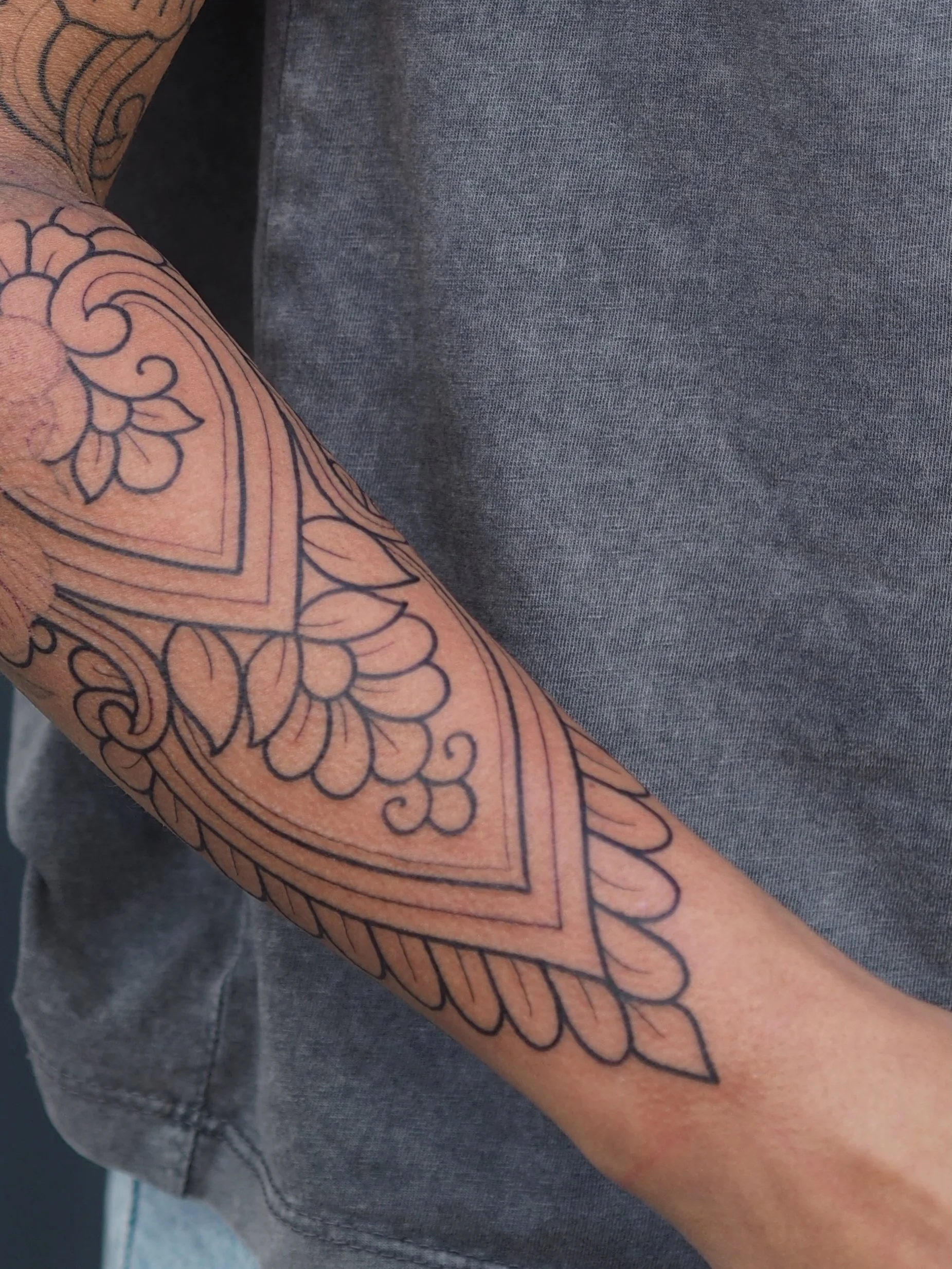 Close-up of a person's arm with a detailed black ornamental outline tattoo featuring floral patterns and decorative border design.