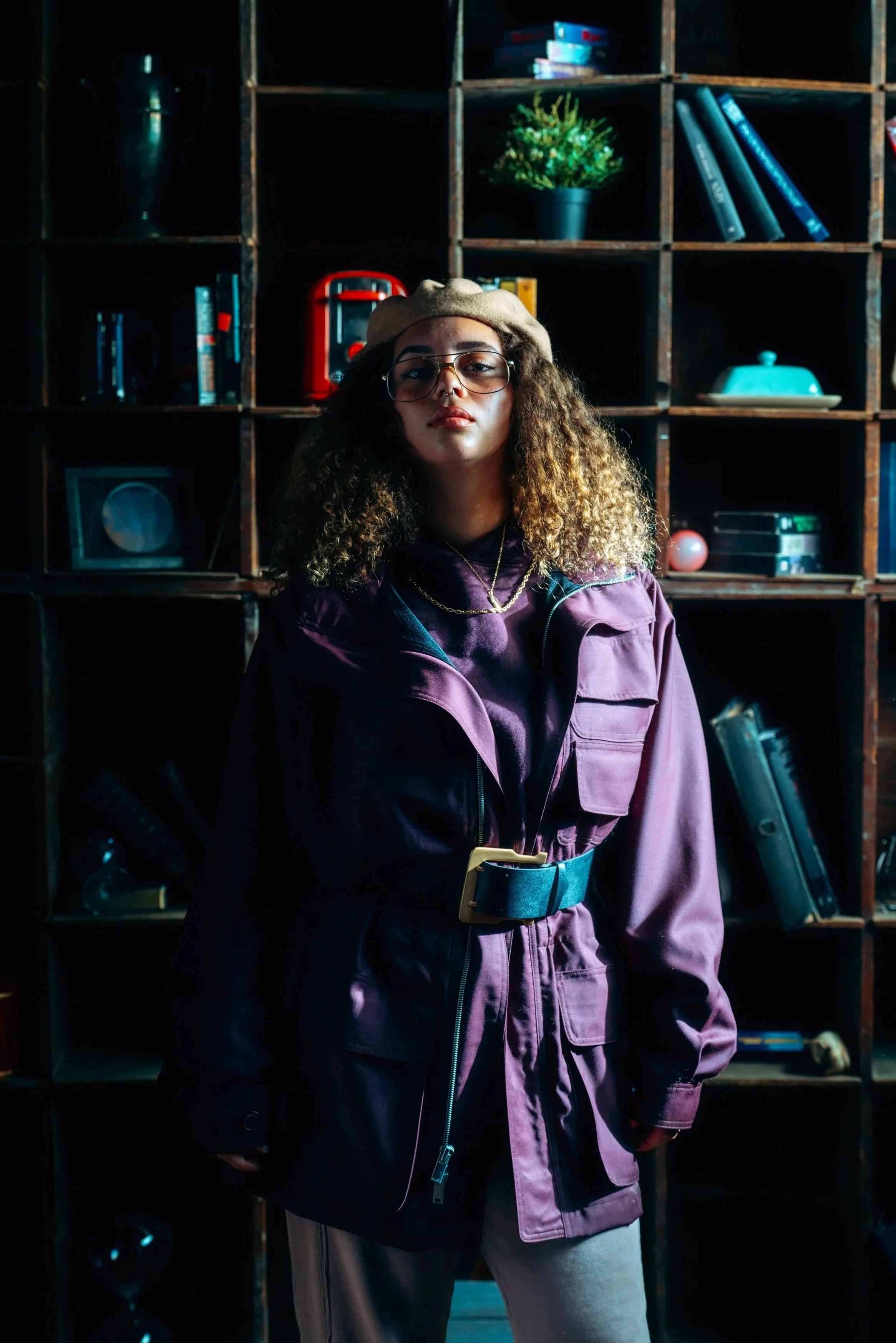 A young woman with curly hair, glasses, and a beret, standing in front of wooden shelves filled with books, a plant, and decorative objects, wearing a purple jacket and gray pants.