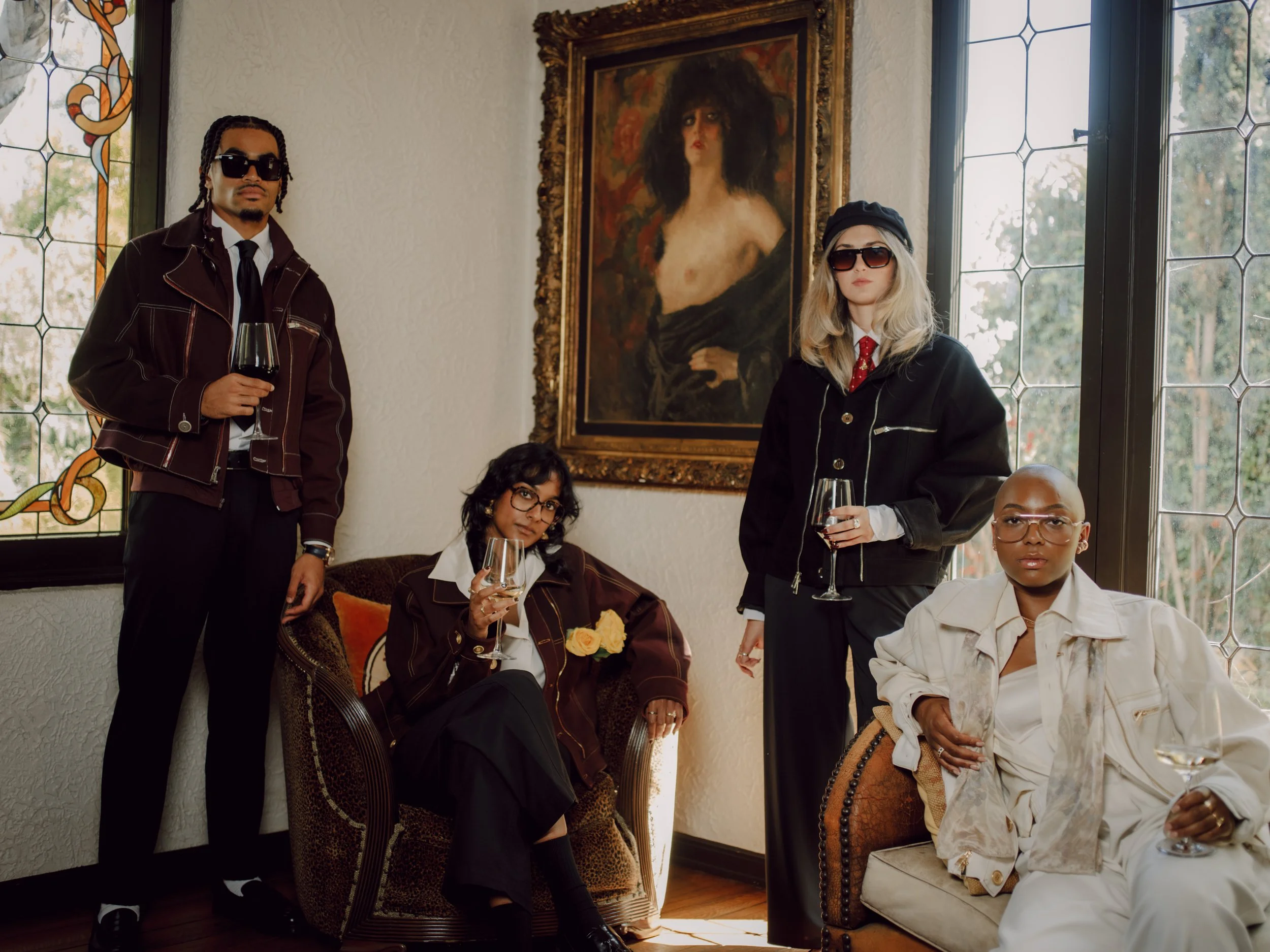 A group of diverse people posing in a room with stained glass windows and a large portrait of a woman on the wall. They are dressed stylishly, some holding wine glasses.