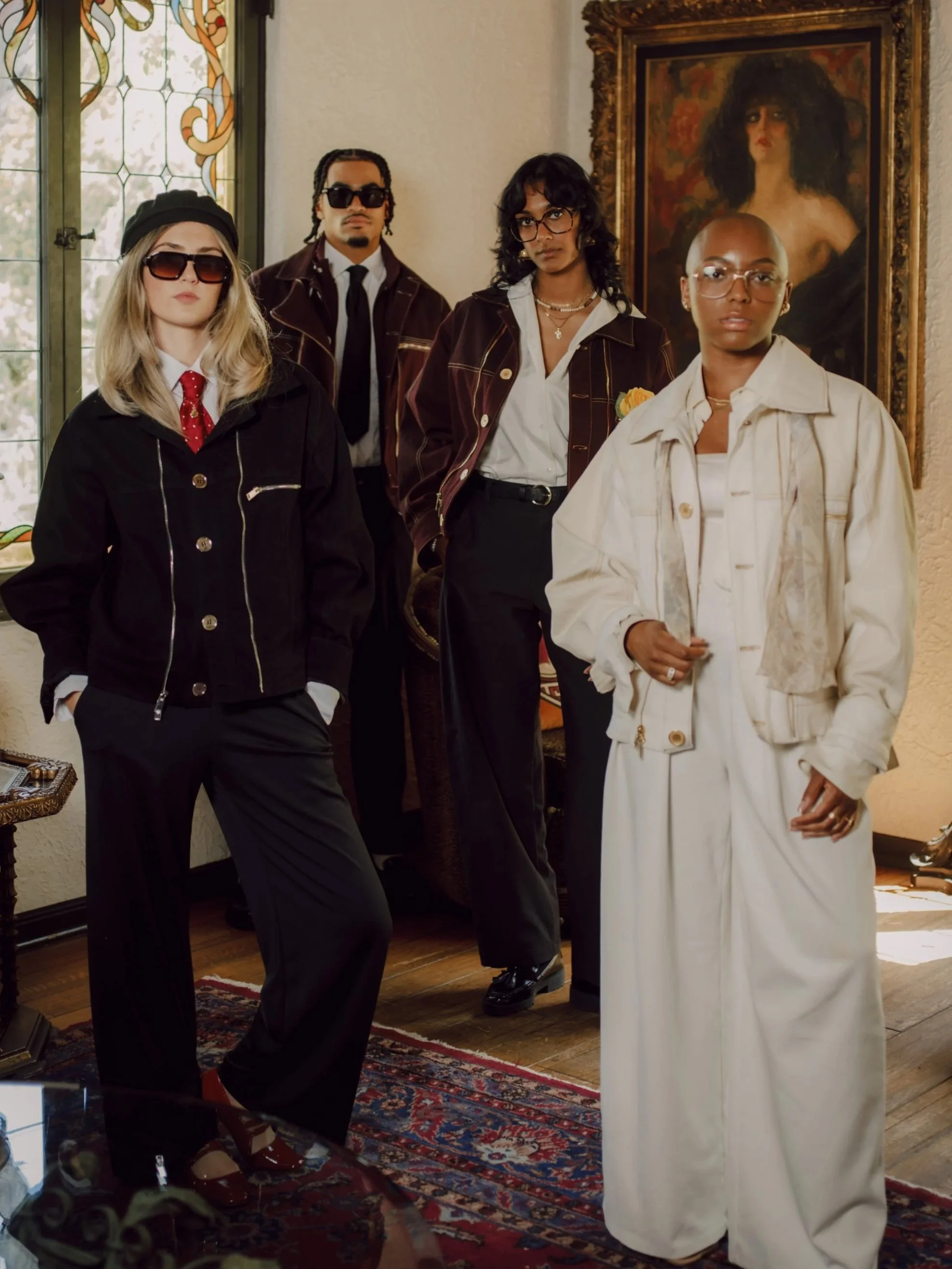Four stylish women with glasses in a room with a large framed painting of a woman with black hair and a flowering background. The women are dressed in fashionable outfits, with one in white, one in black, and two in dark brown jackets.