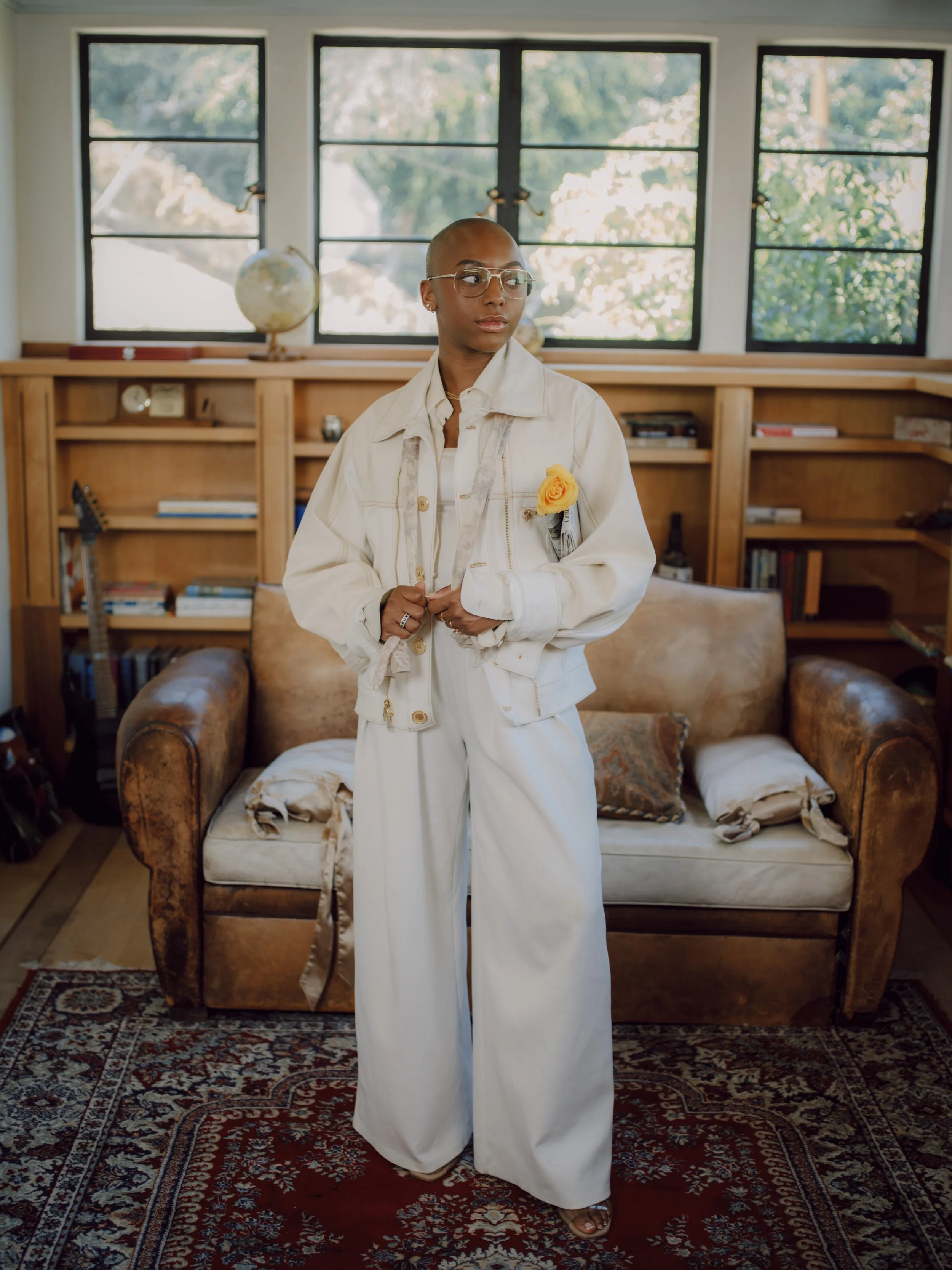 A person with a shaved head and glasses stands in a room with large windows, wearing an oversized white jacket with a yellow flower pinned on it and white wide-leg pants.