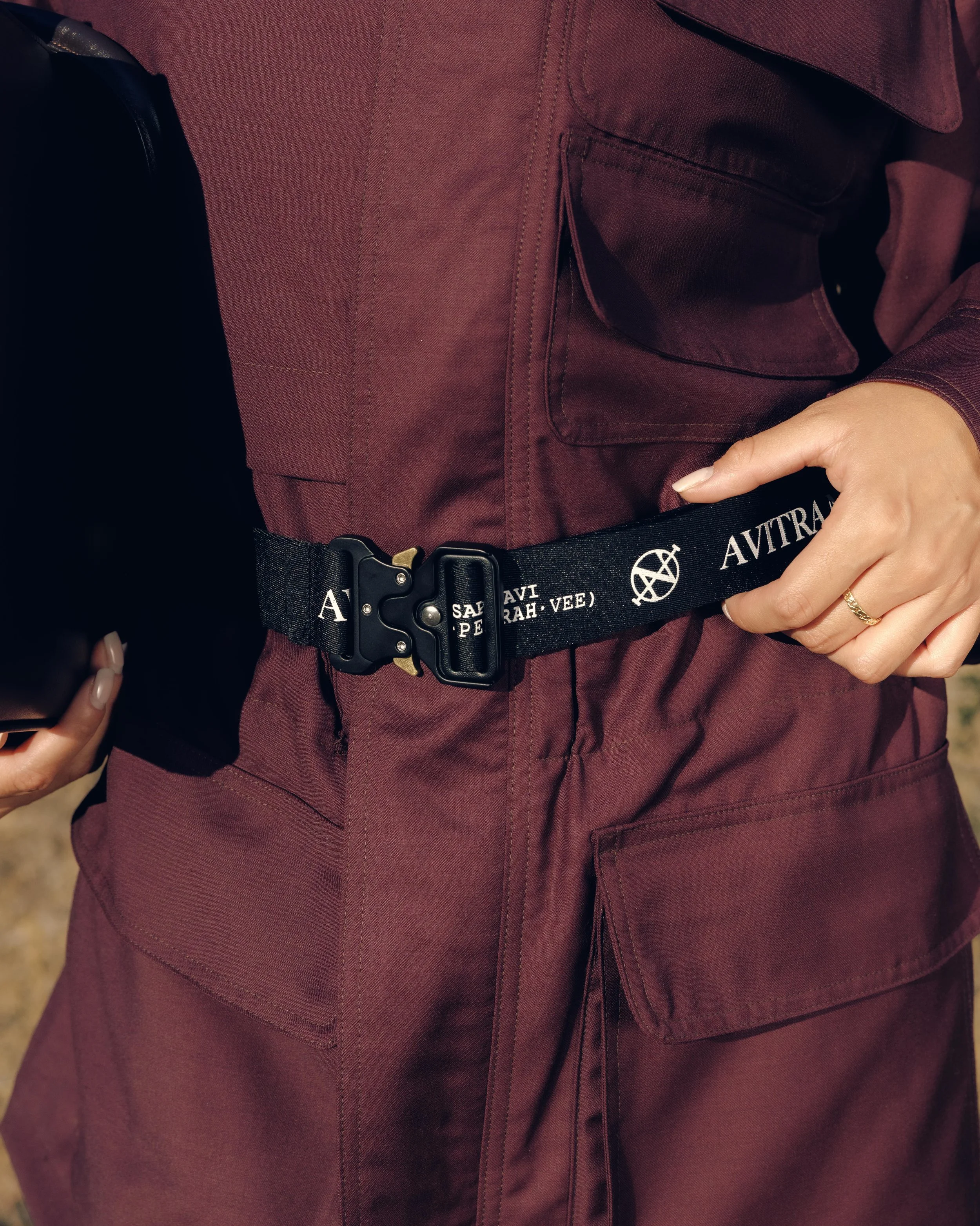 Close-up of a person wearing a maroon jacket with a large front pocket, a black belt with white text and symbol, and a partially visible gold ring on their finger.