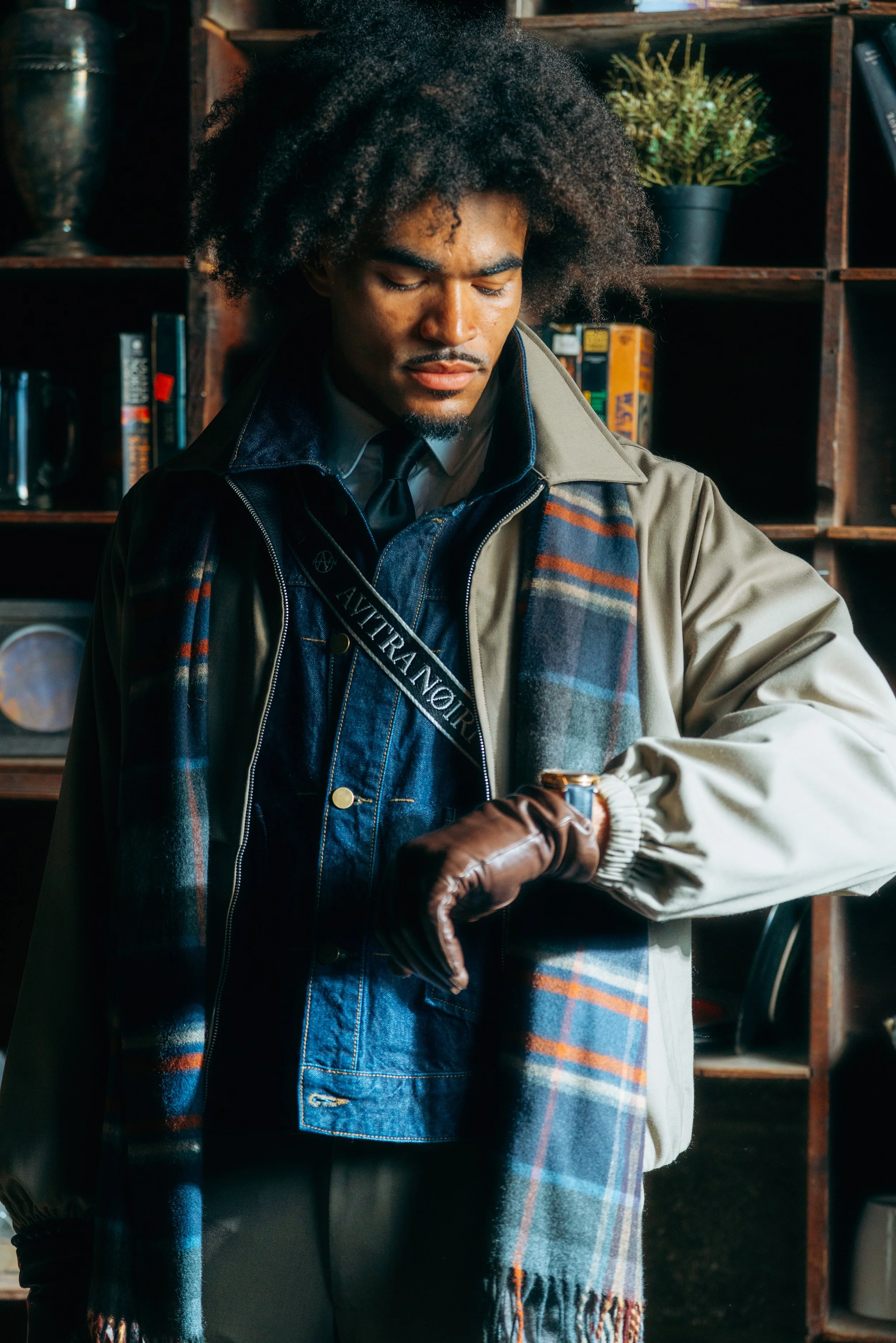 A man with curly black hair wearing a trench coat, denim jacket, and a multicolored plaid scarf, looking at a wristwatch on his left wrist.