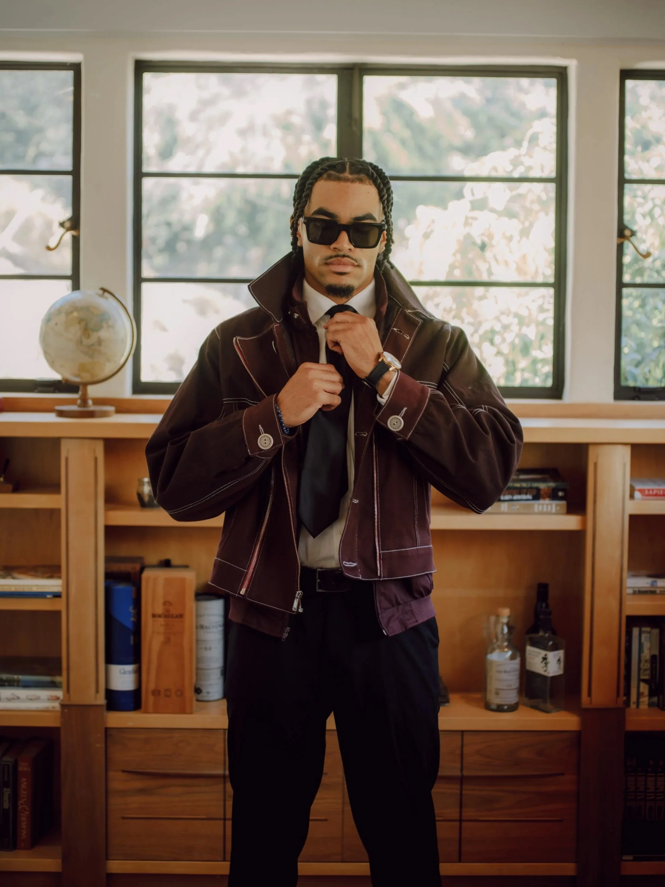 A man with braided hair, wearing sunglasses, a dark jacket, and a watch, standing in front of a wooden bookshelf with books and bottles, inside a room with large windows.