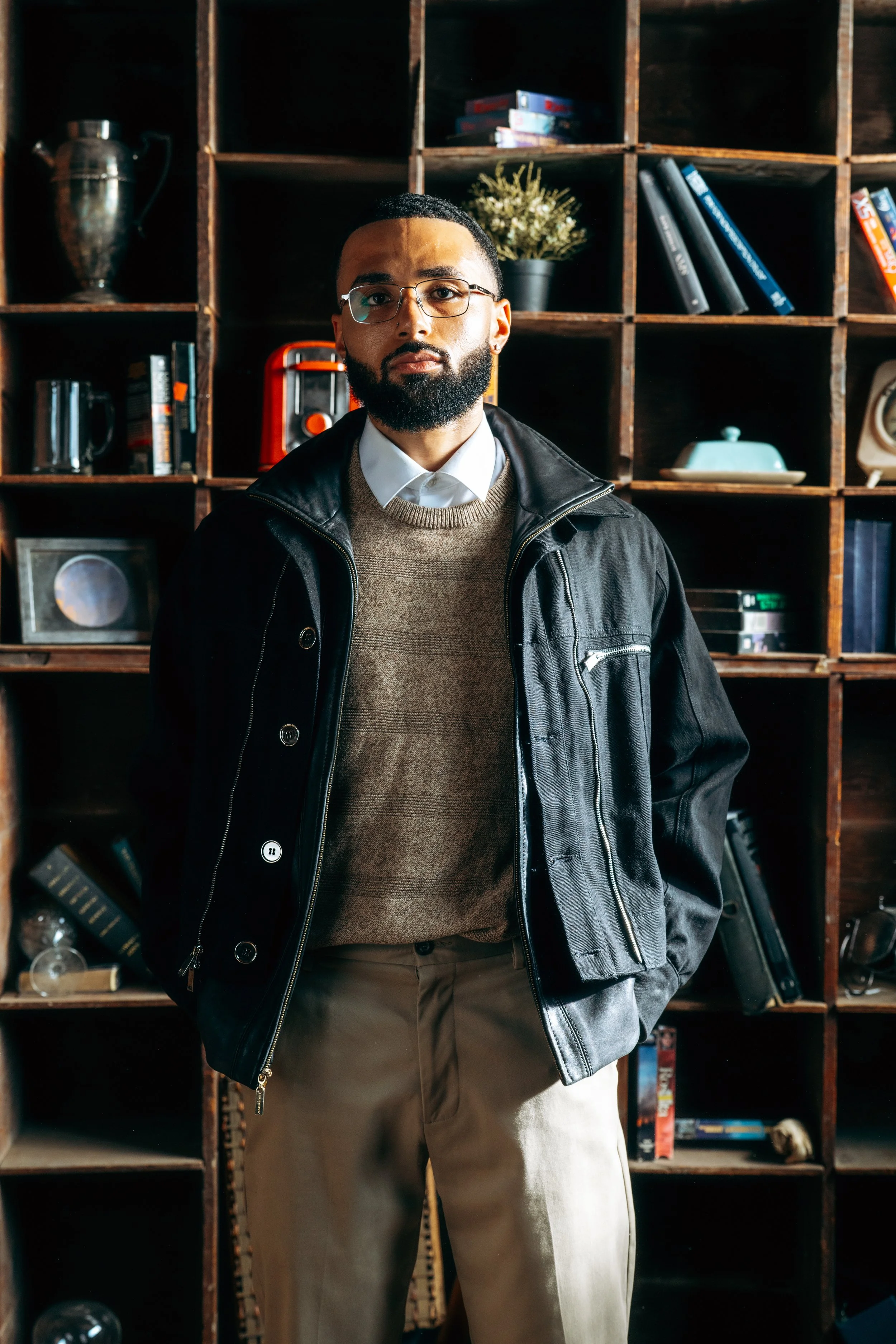 A man with glasses and a beard standing in front of a wooden bookshelf filled with books, a plant, and decorative items.