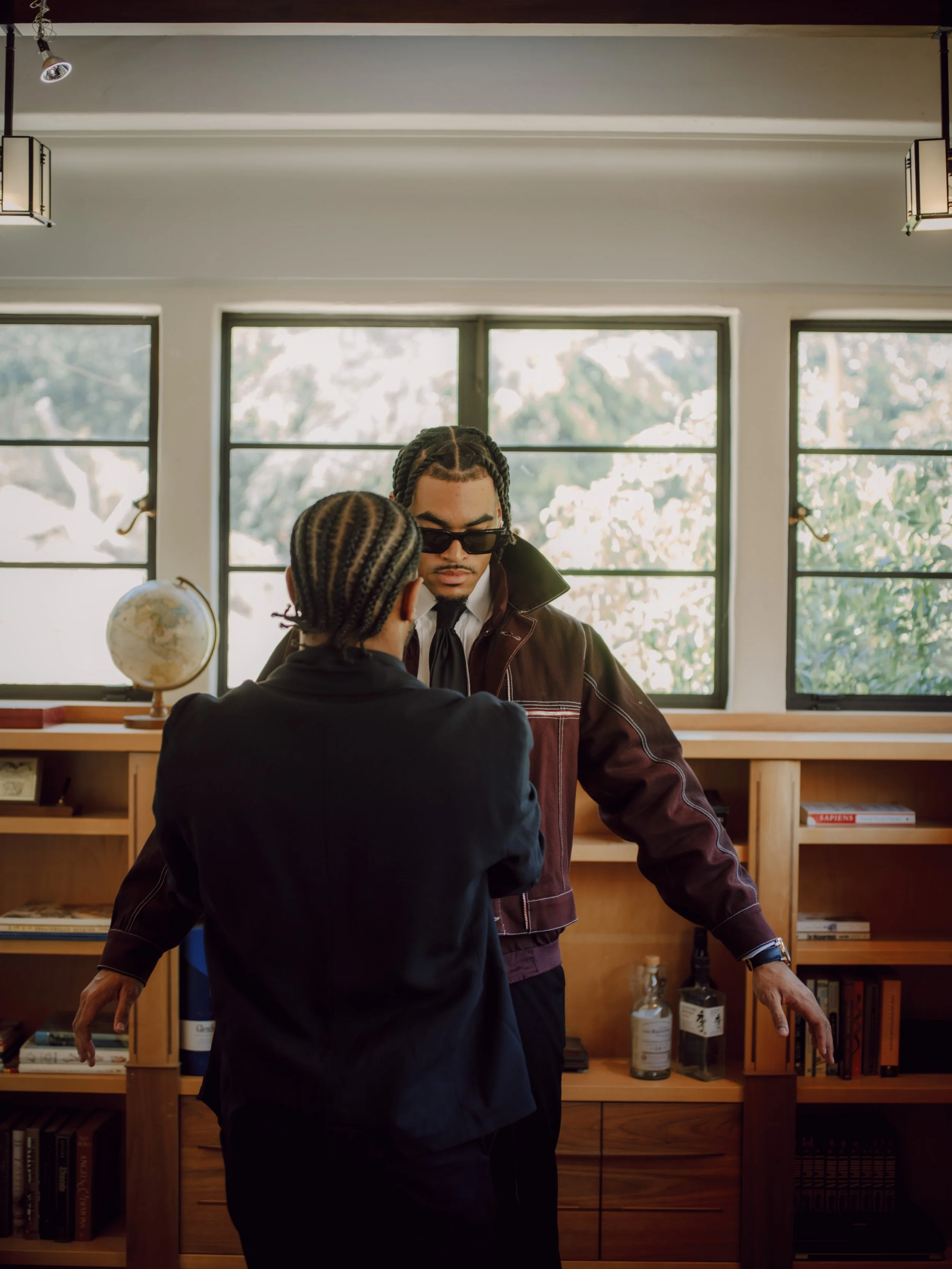 Two men with braided hair are inside a room with large windows, a wooden bookshelf, and a globe. One man wears dark sunglasses and a brown jacket, while the other faces away and wears a black hoodie.