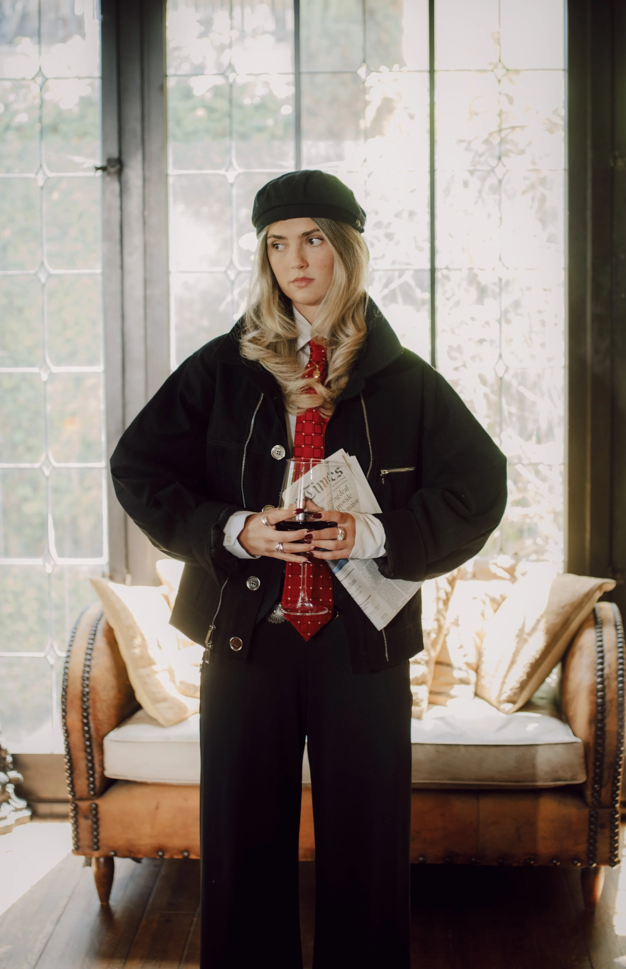 Woman with blonde hair wearing a black beret, black jacket, and red tie, holding a glass of wine and newspapers, standing in front of a large window with natural light.