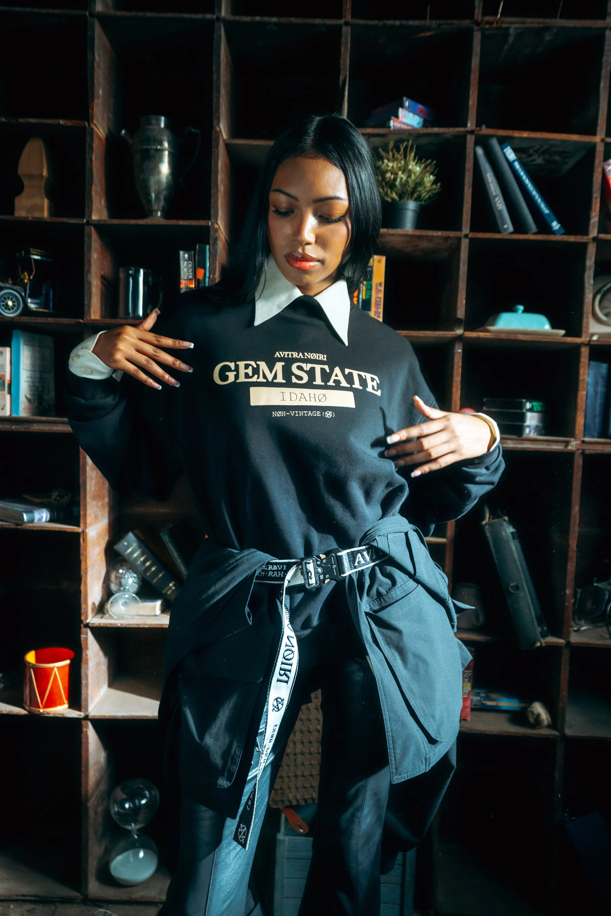 A woman with dark hair wearing a black sweatshirt with white text and black pants with a white belt stands in front of wood shelving filled with various objects, including books, plants, and decorative items.