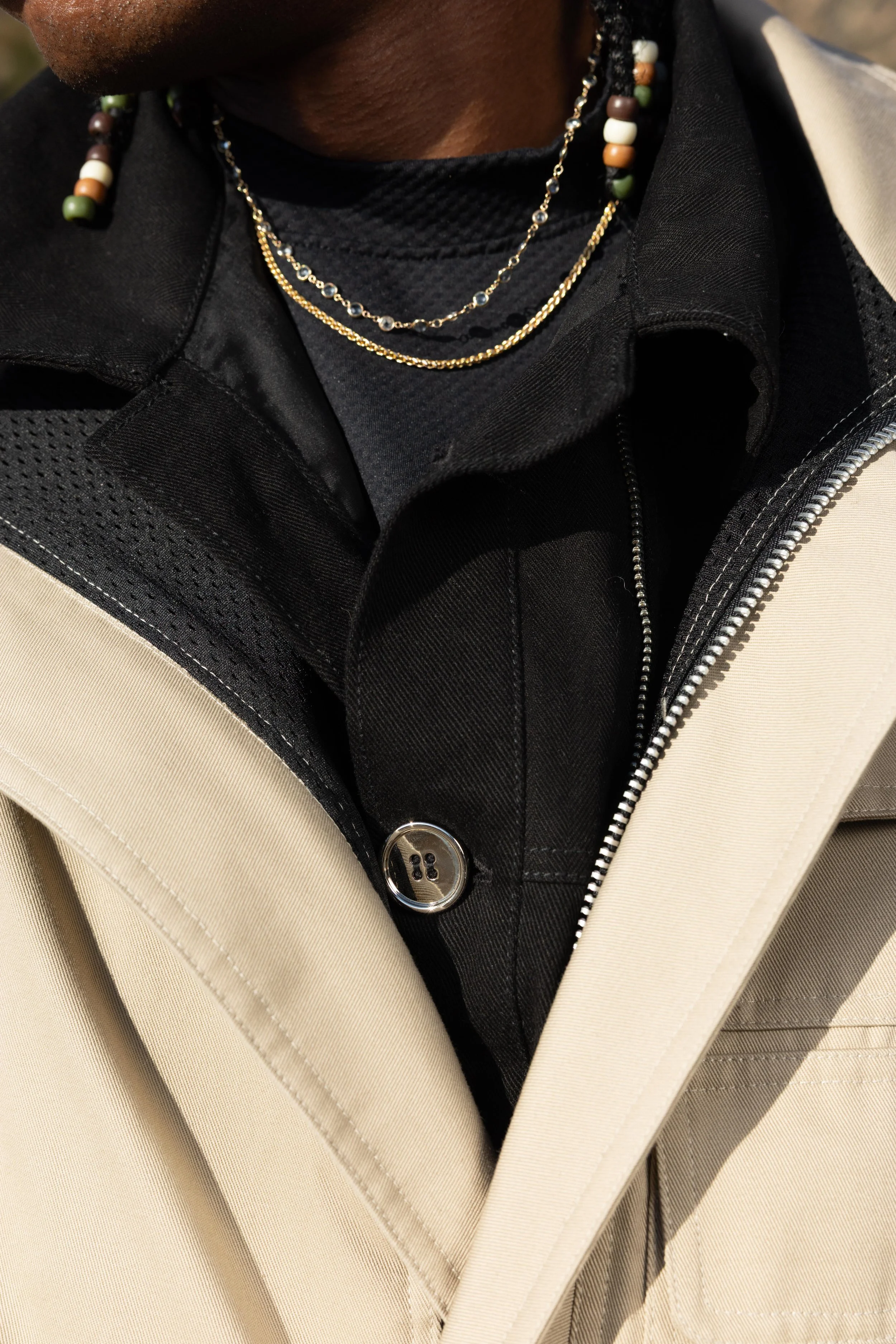 Close-up of a person's neck wearing layered necklaces, black shirt, and beige jacket with a zipper.