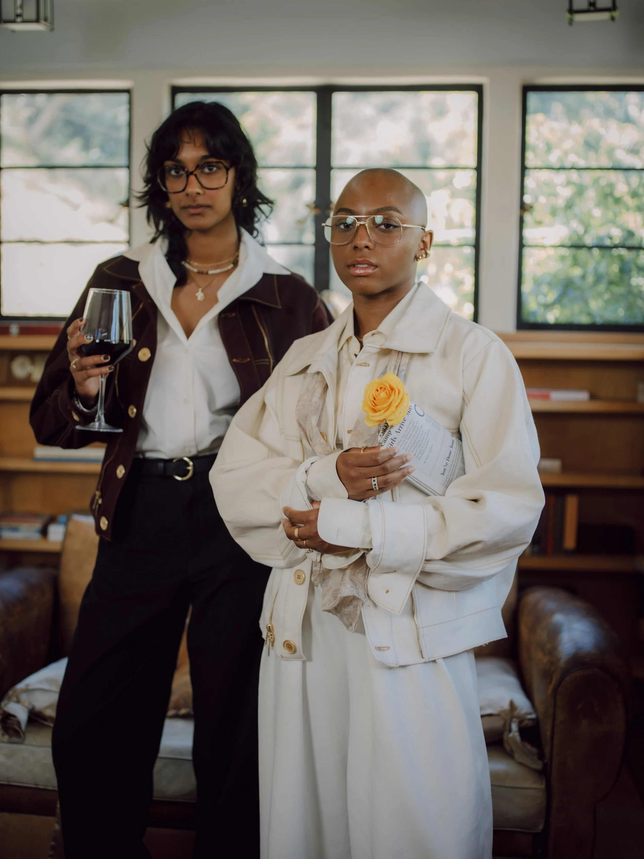 Two women with glasses standing in a living room, one holding a glass of red wine, both looking at the camera.