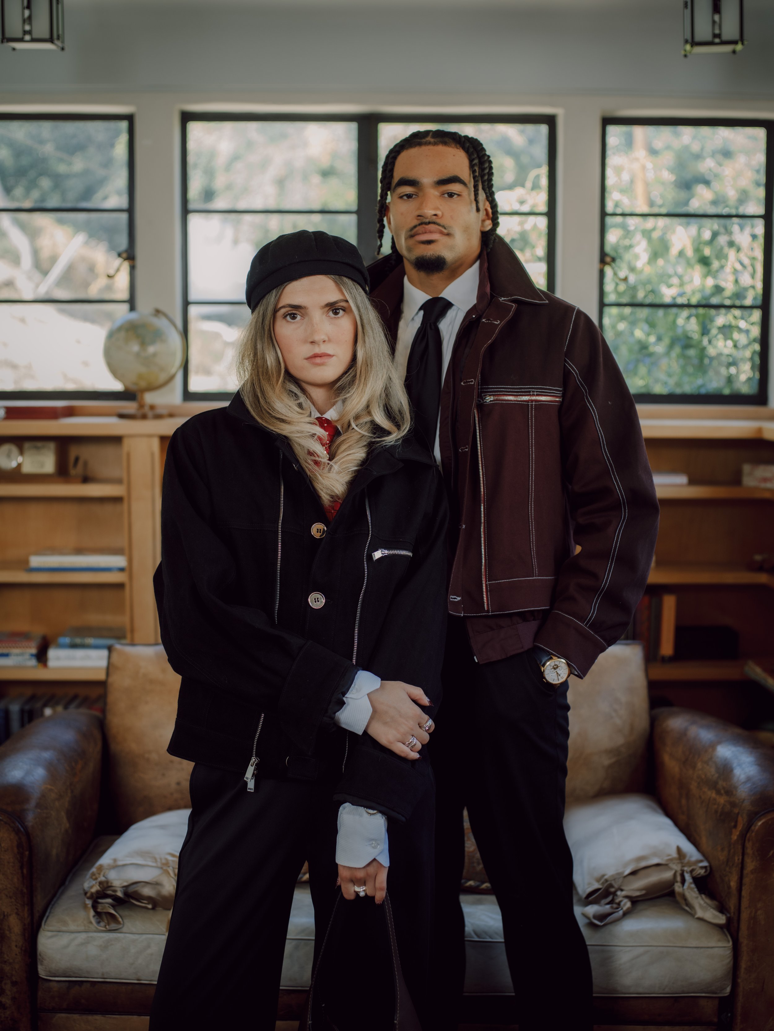 A man and a woman standing close together inside a room with large windows and a bookshelf in the background. The woman has long blonde hair, light skin, and is wearing a black hat and a black jacket. The man has dark skin, braided hair, and is dress
